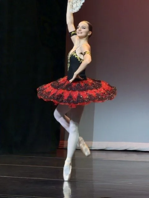 Ballet dancer in a black and red tutu performing on stage, standing on one leg, with a large white feathered prop behind her.