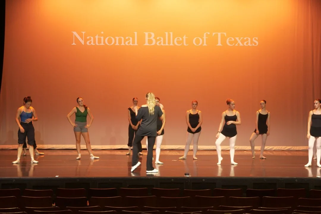 Group of ballet dancers practicing on stage with a director, in front of a backdrop that reads 'National Ballet of Texas'.