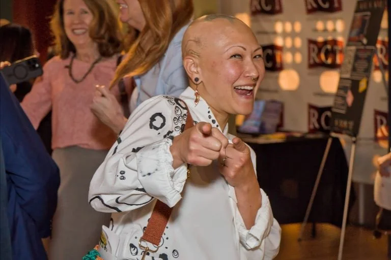A cheerful woman with a shaved head pointing at the camera, smiling, and surrounded by people at a social event.
