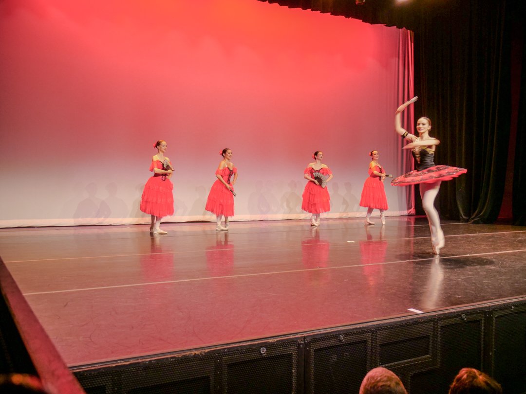 Ballet dancers performing on stage, with one dancer in front wearing a black and red tutu and white tights, and the other four dancers in red dresses holding black fans, performing a ballet routine.