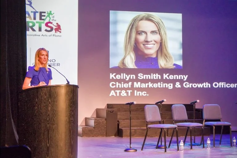 Woman in a blue dress speaking at a wooden podium on stage with chairs and microphones, projected slide shows her photo and name Kellyn Smith Kenny, Chief Marketing & Growth Officer at AT&T Inc., during an event with a banner reading 'Creative Arts o