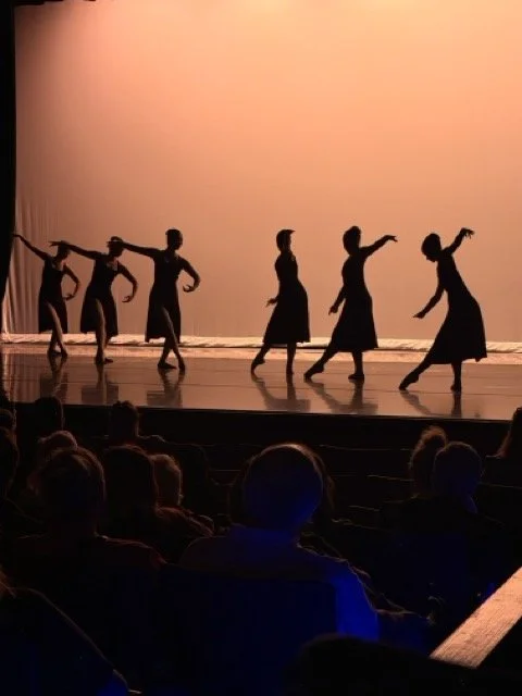 Silhouettes of six dancers performing on stage with an audience watching in the foreground.