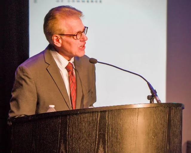 A man in a gray suit, white shirt, and red striped tie speaking at a podium with a microphone, wearing glasses and with hair slicked back.