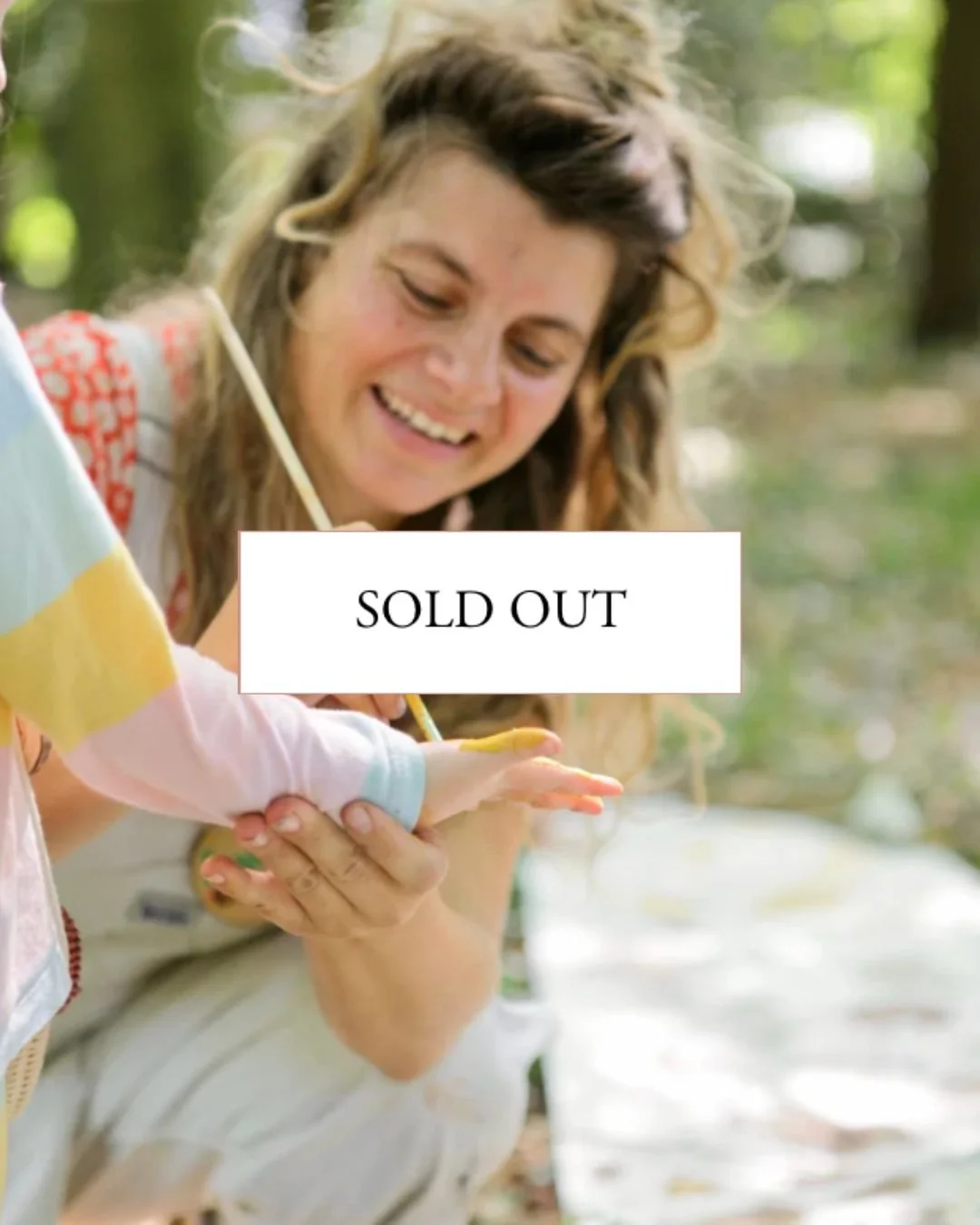Woman smiling while painting a child's hand in a park or forest setting with a white cloth on the ground.