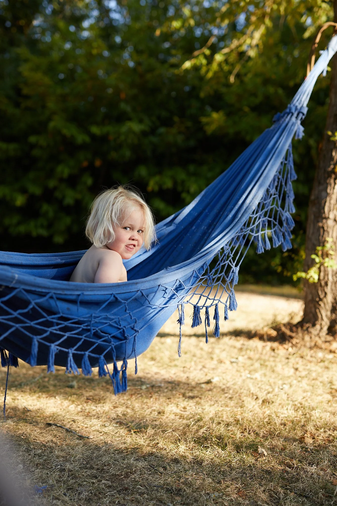 A young child with blonde hair and a mischievous expression lying in a blue hammock tied to a tree outside, surrounded by green foliage.