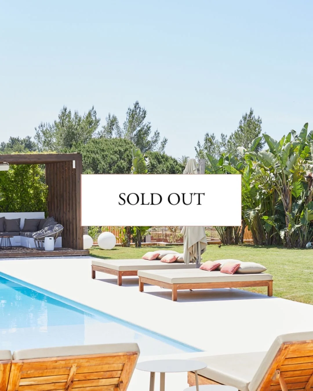 A backyard swimming pool area with wooden lounge chairs, beige cushions, and a white side table, surrounded by lush green trees and plants on a sunny day.