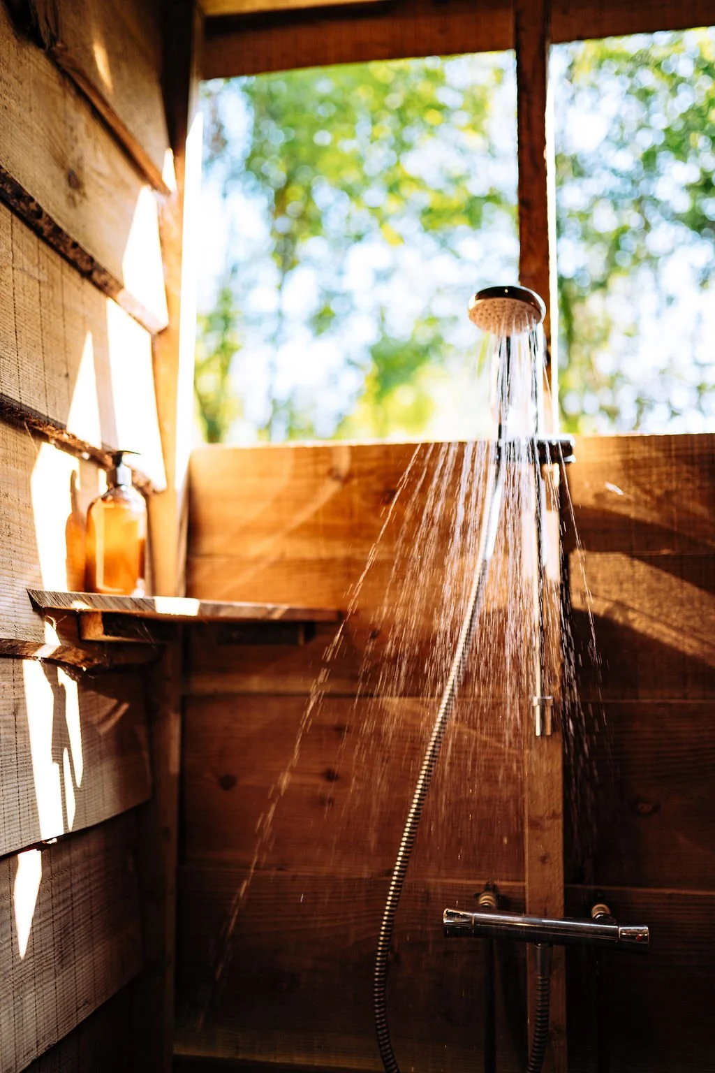 An outdoor shower with water flowing from a round showerhead, mounted on a wall made of warm-colored wooden panels, with a soap dispenser on a small ledge nearby, and green trees in the background.