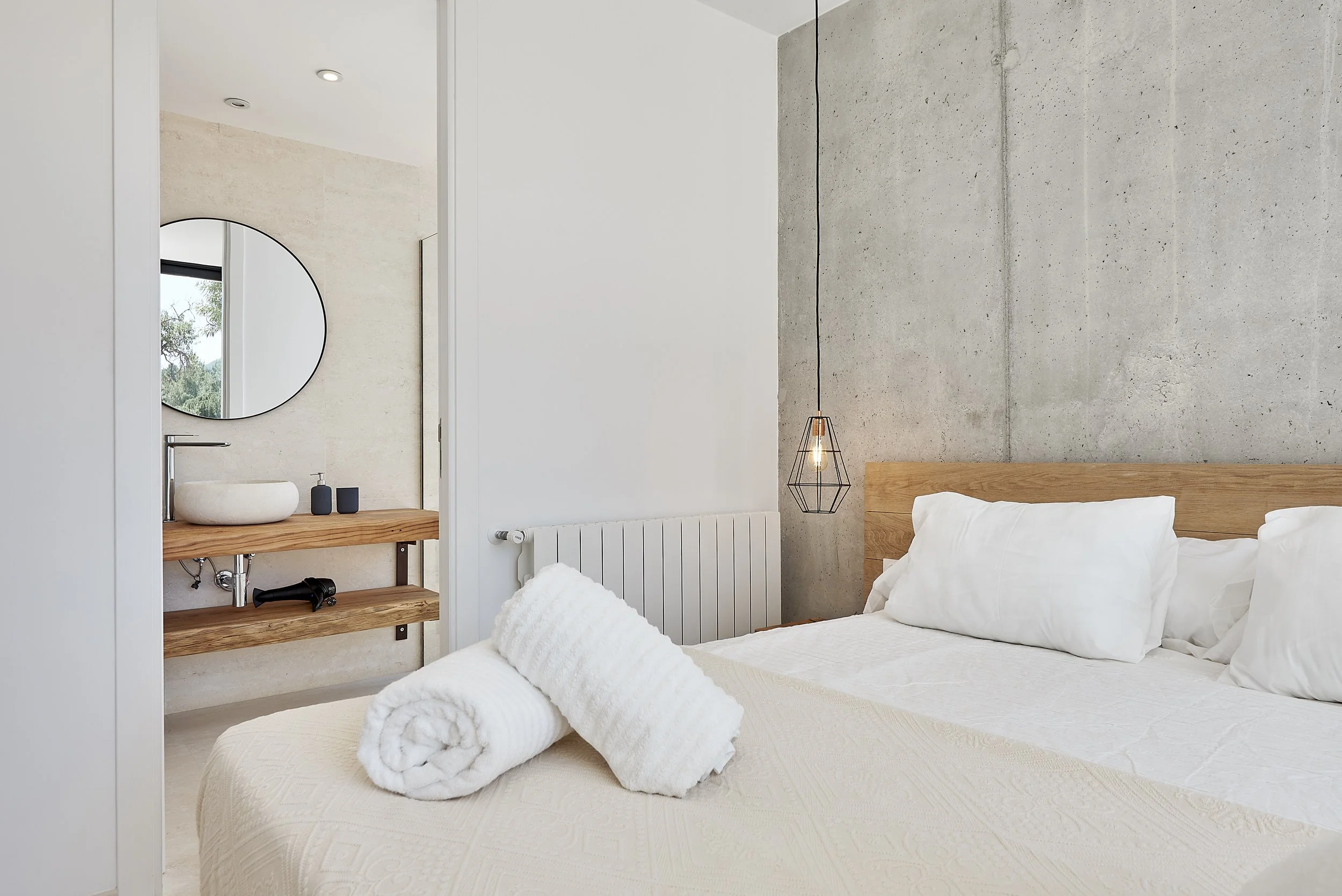 Modern bedroom with a bed, white pillows, and rolled towels. A concrete accent wall with a hanging pendant light. An adjacent area with a wooden sink and mirror, seen through a partially open door.
