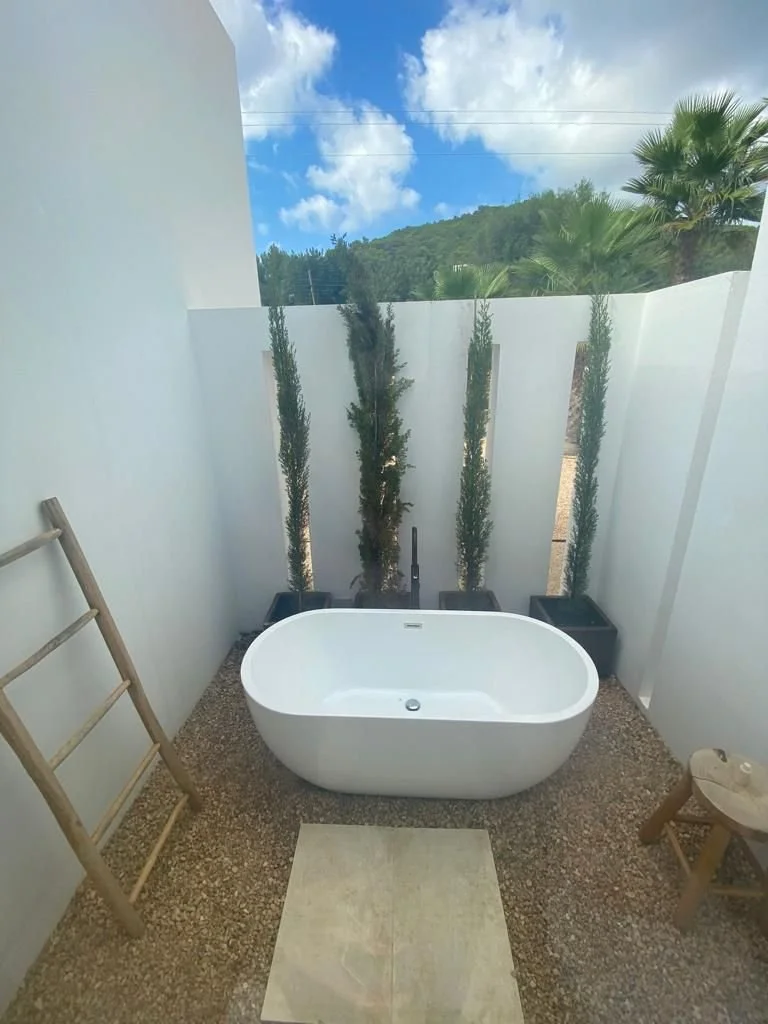 Outdoor bathtub area with white tub, potted tall, narrow trees, wooden ladder, and small stool, against a backdrop of green hills and blue sky with clouds.
