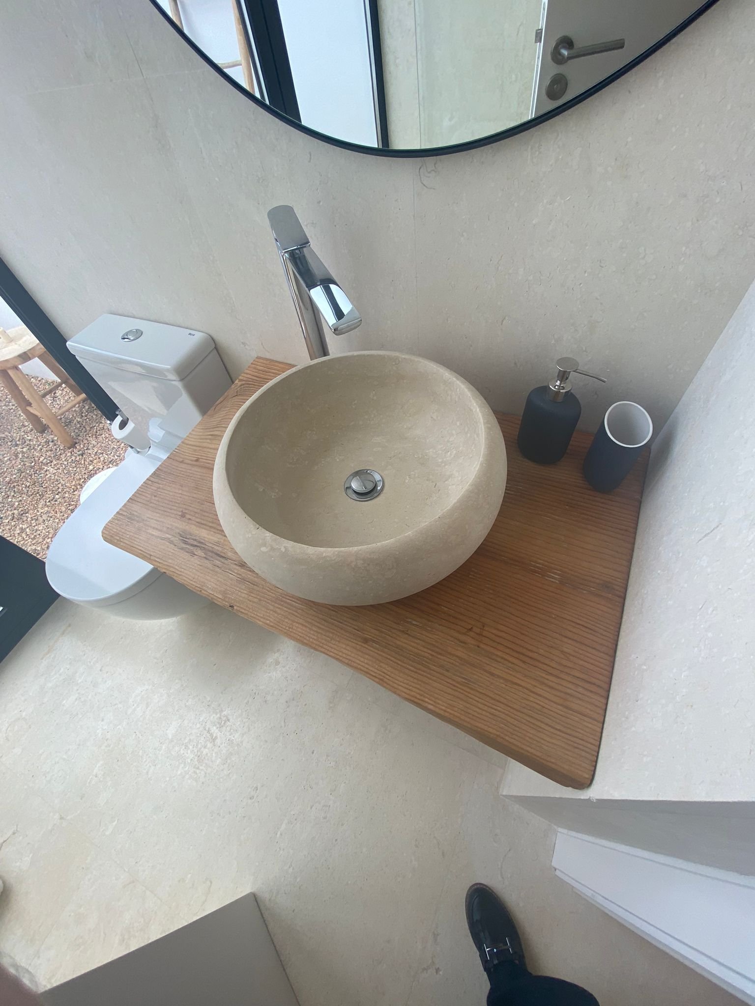 A bathroom sink made of stone on a wooden countertop with a mirror above. A soap dispenser, a tumbler, and a faucet are also on the countertop. A toilet is visible in the background.