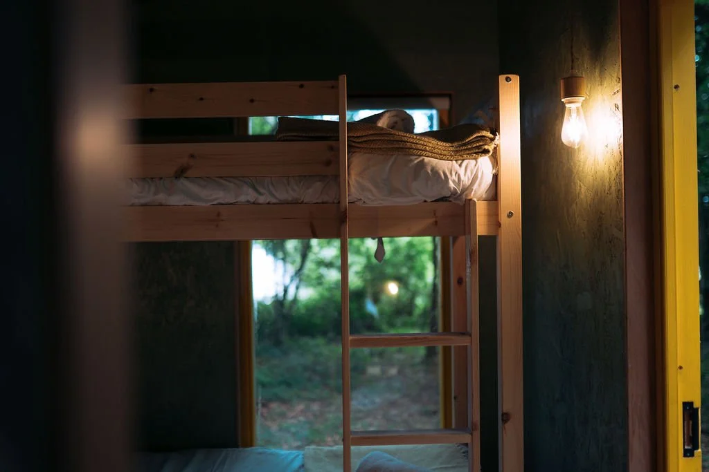 Interior of a cozy bedroom with a wooden bunk bed, a small window showing greenery outside, and a warm wall-mounted light bulb.