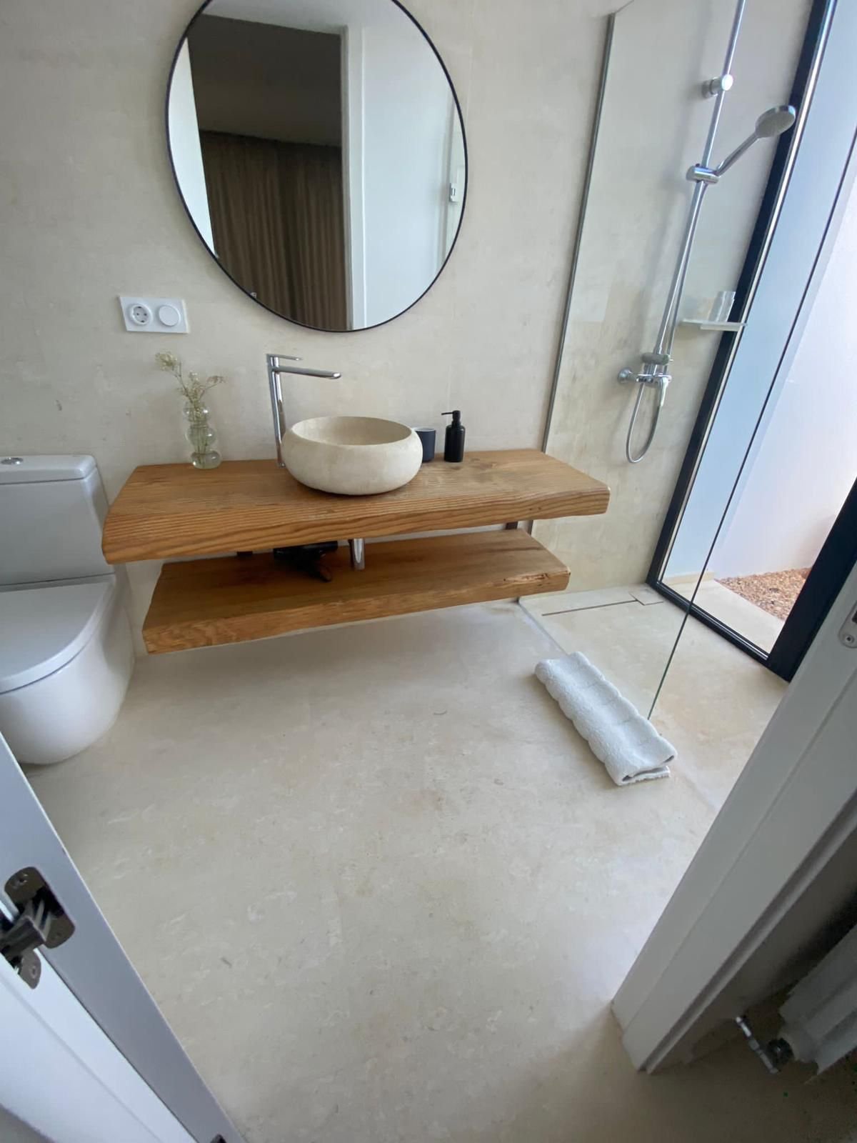 Modern bathroom with a round mirror, stone vessel sink on a wooden countertop, black soap dispenser, rolled white towel on the floor, and a walk-in shower with a glass door and a rain shower head.