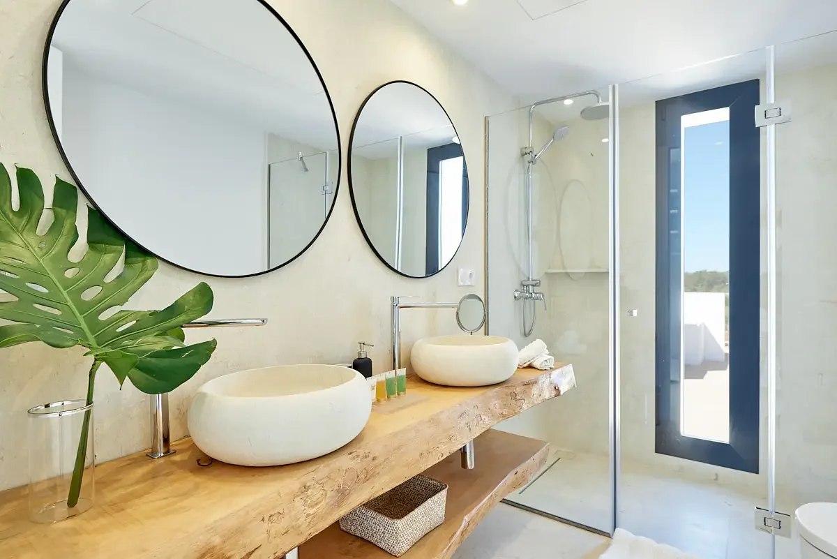 Modern bathroom with two round mirrors over a wooden countertop with two vessel sinks, a shower area with glass door, and a window letting in natural light.