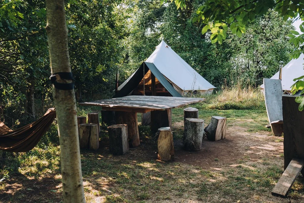 A camping scene with a circular wooden table surrounded by log stools, a white canvas tent in the background, and trees providing shade.
