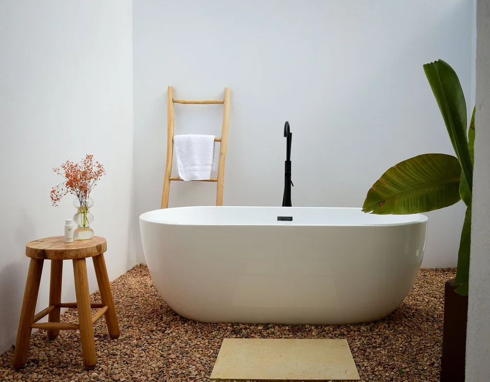 Modern bathroom with a white bathtub, a wooden ladder with a towel, a small wooden stool with toiletries, large green plants, and a pebble stone floor.