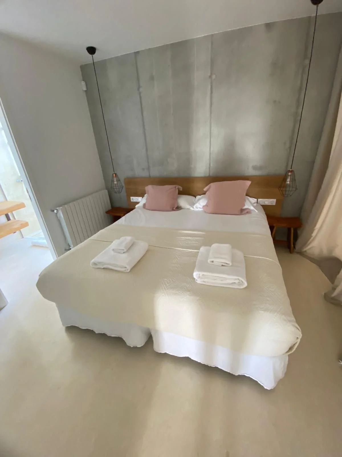 A neatly made hotel bed with two pink pillows, two sets of folded white towels, and a cream-colored bedspread. There are two wooden nightstands on either side with hanging pendant lights above them. The room has a concrete accent wall and a sliding glass door partially visible on the left.