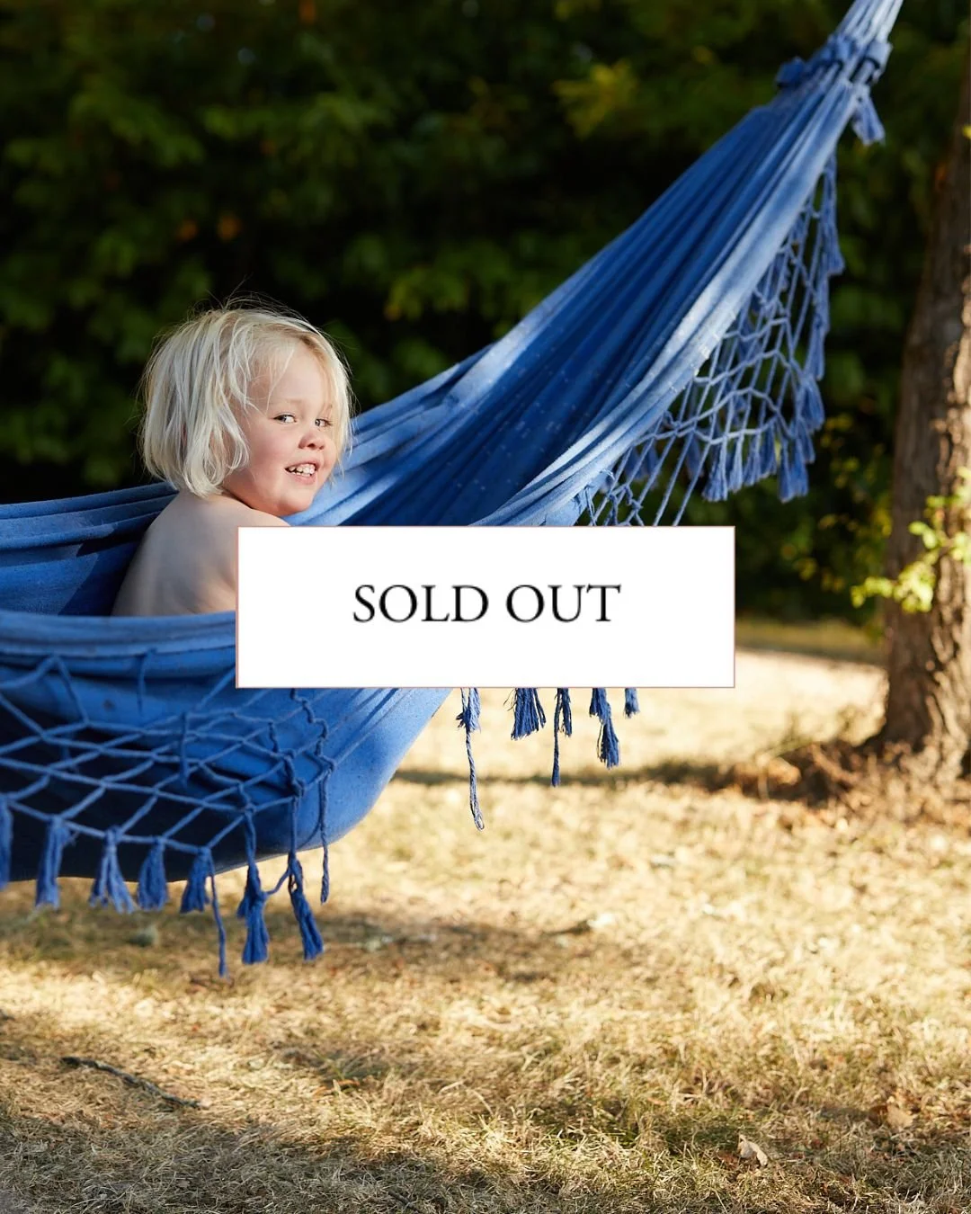 A young child with blonde hair and a mischievous expression lying in a blue hammock tied to a tree outside, surrounded by green foliage.