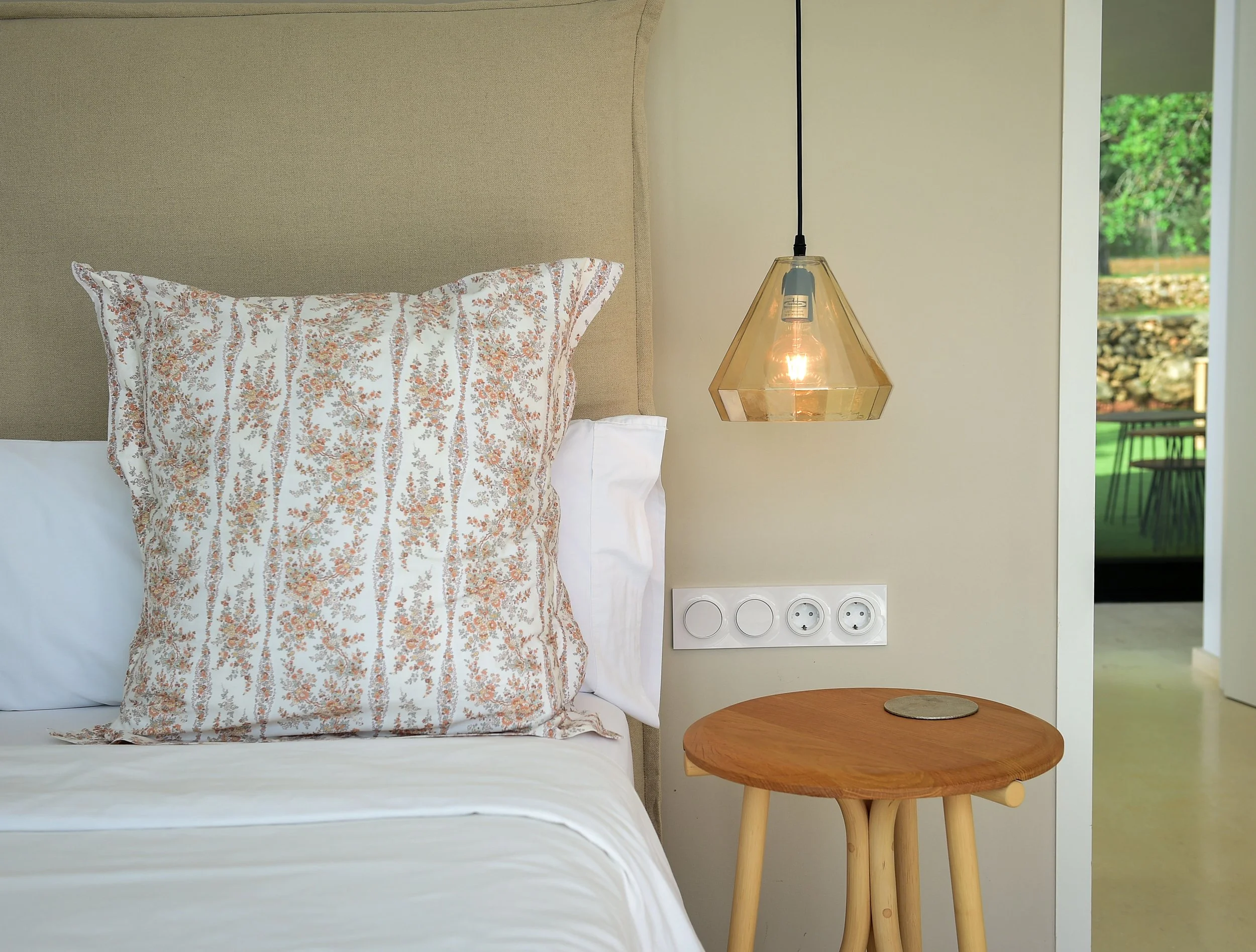 A bed with a beige headboard, a white pillow, and decorative pillow with a floral pattern, a hanging pendant light, a wooden side table, and a wall with electrical outlets. There is a door open to a balcony with outdoor furniture and green trees outside.