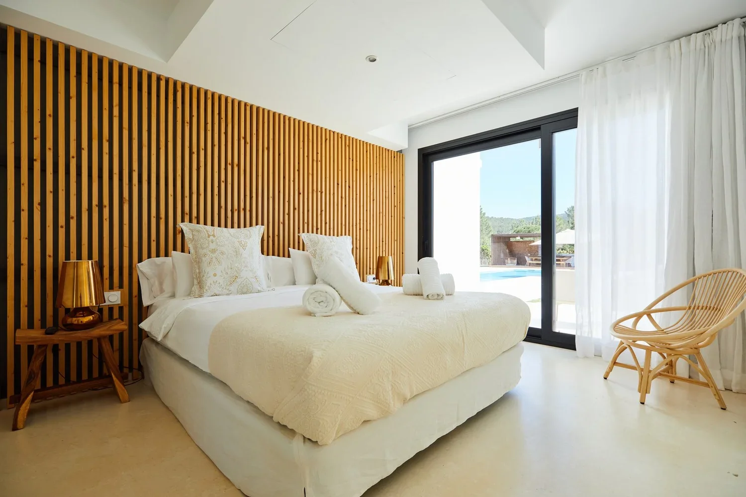 A bedroom with a large bed covered in cream-colored bedding and decorative pillows, facing a sliding glass door with white curtains. There are rolled towels on the bed, a small wooden side table with a copper lamp, and a rattan chair near the window. The wooden accent wall behind the bed adds warmth to the minimalist decor, and outside is a view of a pool and trees.