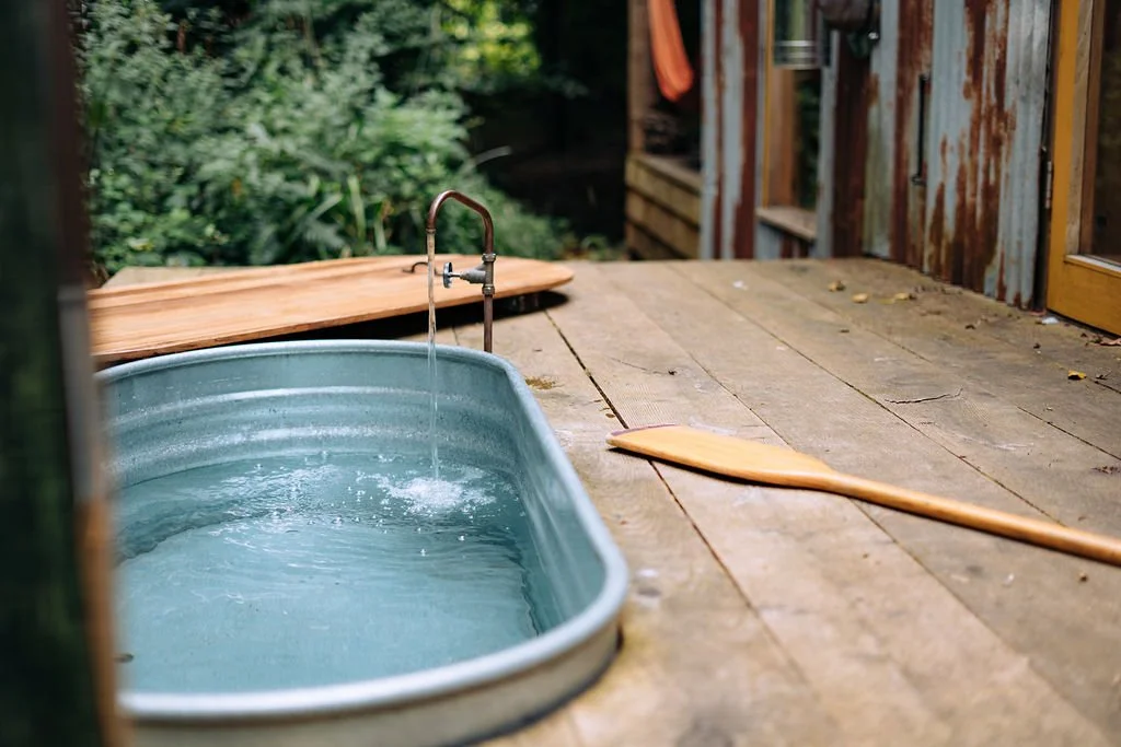 An outdoor wooden deck with a small greenish metal tub filled with water, a wooden spoon resting on the deck, and a faucet with water running into the tub.