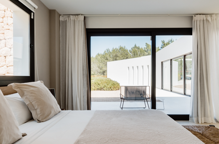 Bedroom with large sliding glass door opening to patio with chair, outdoor areas, and trees in background.