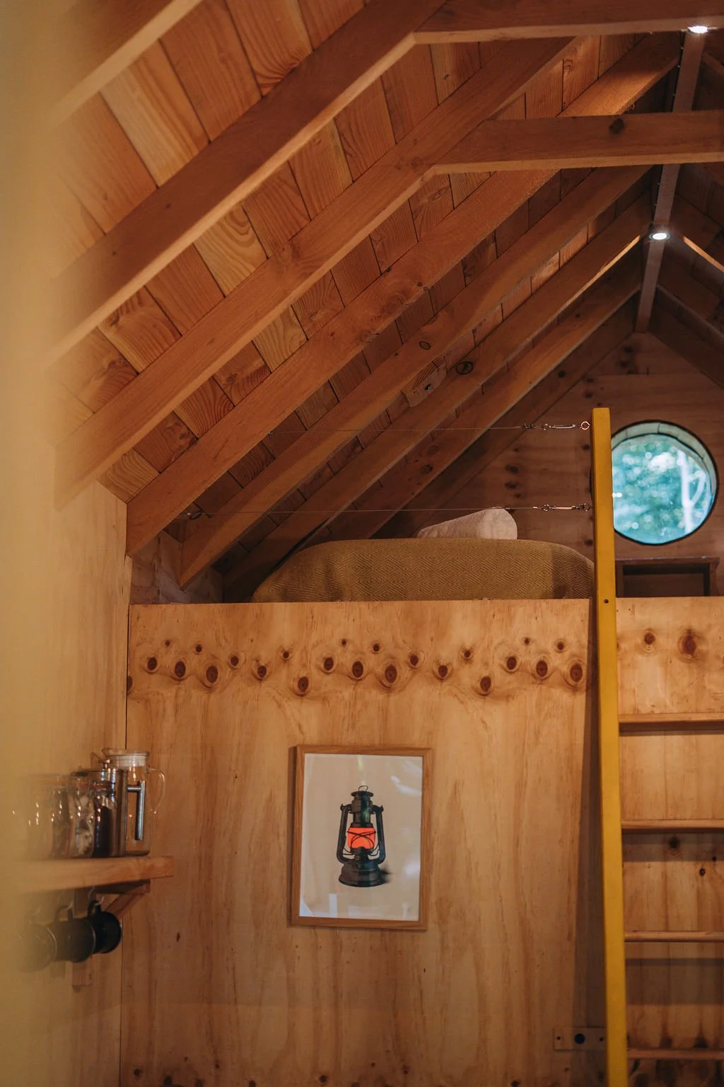 The interior of a wooden cabin with a loft area. The ceiling is made of angled wooden planks supported by wooden beams. In the lower part of the image, there is a small framed picture of a lantern hanging on a wooden wall, a yellow ladder leading to the loft, and a bed with a pillow next to a round window showing trees outside. On the left, a small shelf holds glass jars, and a lantern painting hangs on the wall.