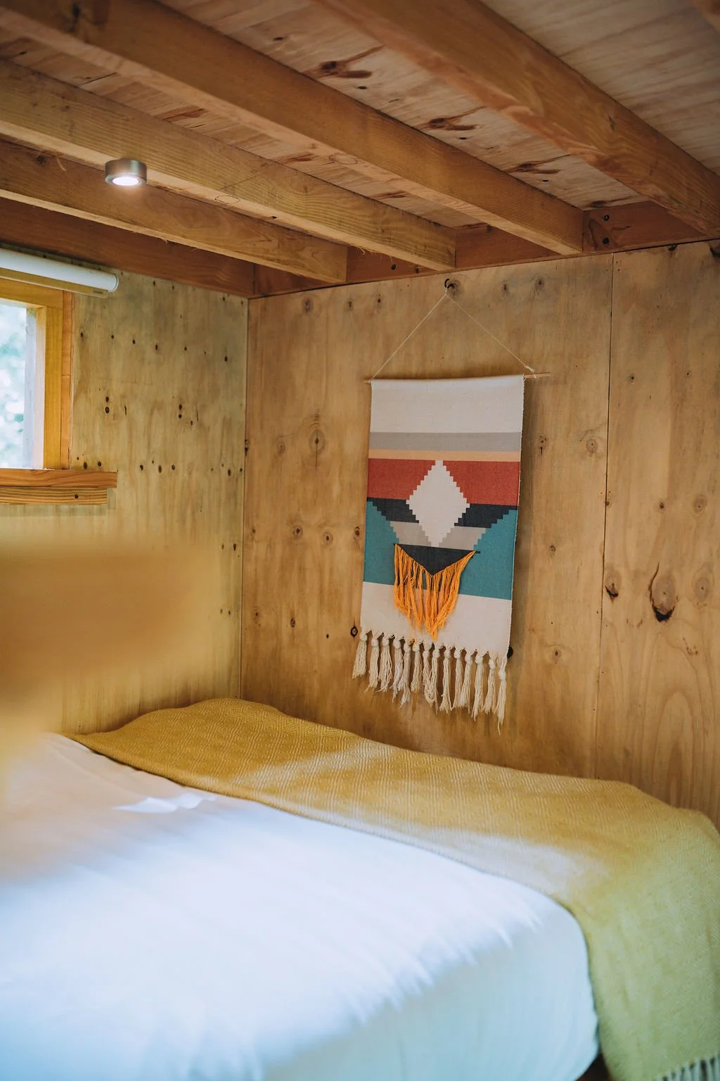A cozy bedroom with wooden walls and ceiling, a small window, a bed with white sheets and a yellow blanket, and a colorful woven wall hanging with tassels.