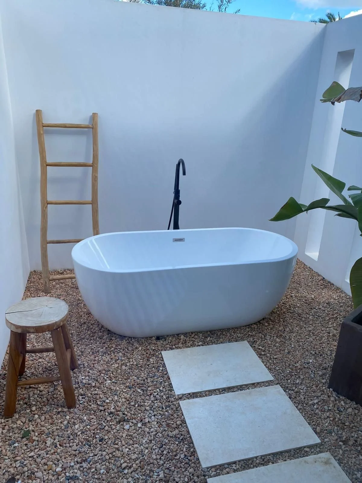 Outdoor freestanding bathtub with a black faucet, wooden ladder, small wooden stool, and stepping stones on gravel, enclosed by white walls with a small window on the right.