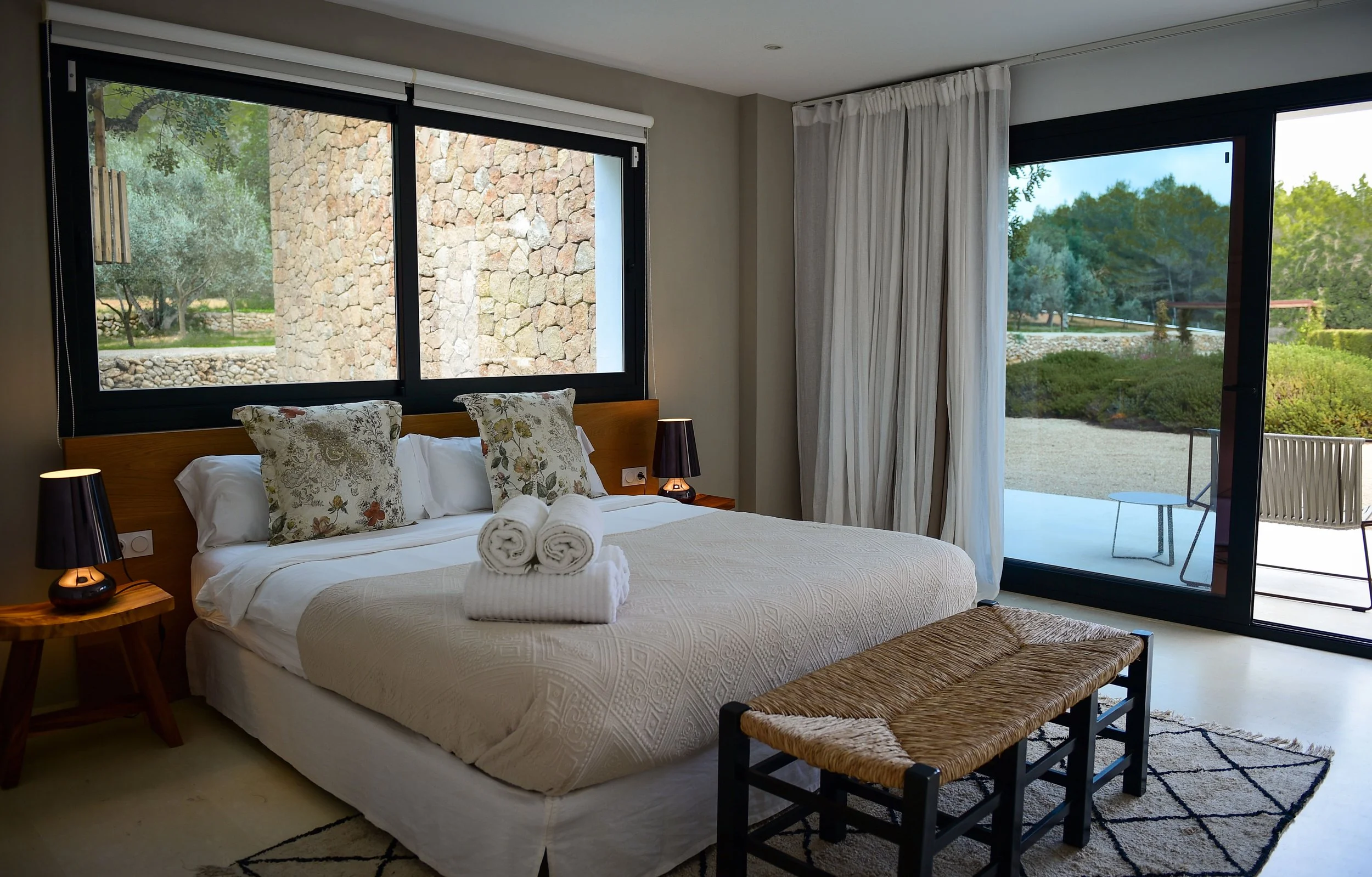 A bedroom with a bed, decorative pillows, rolled towels, bedside lamps, large windows, and a sliding glass door leading to an outdoor patio with outdoor furniture.