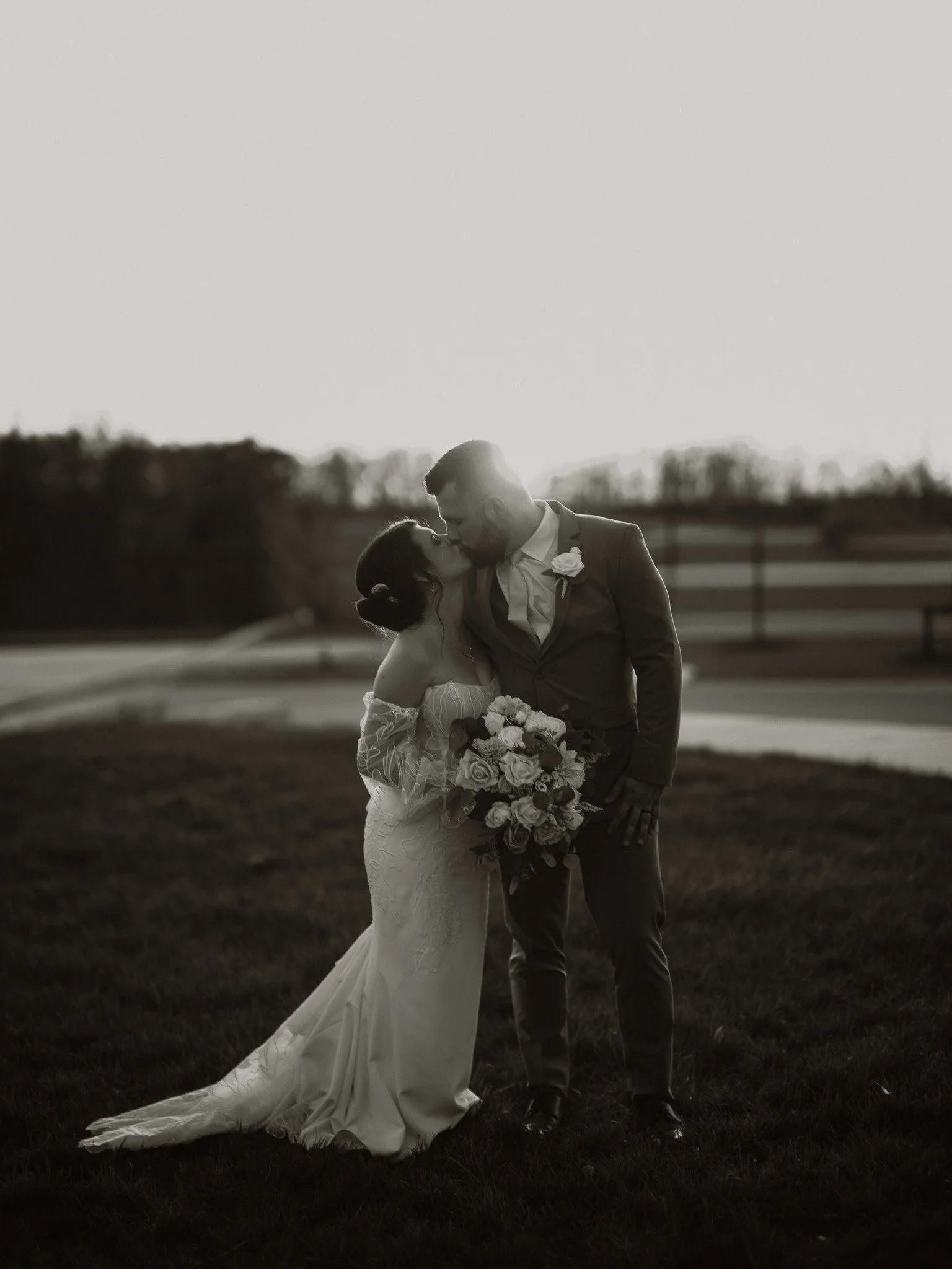 A bride and groom share a kiss outdoors at sunset, with the bride holding a bouquet of flowers and wearing an off-shoulder wedding dress.