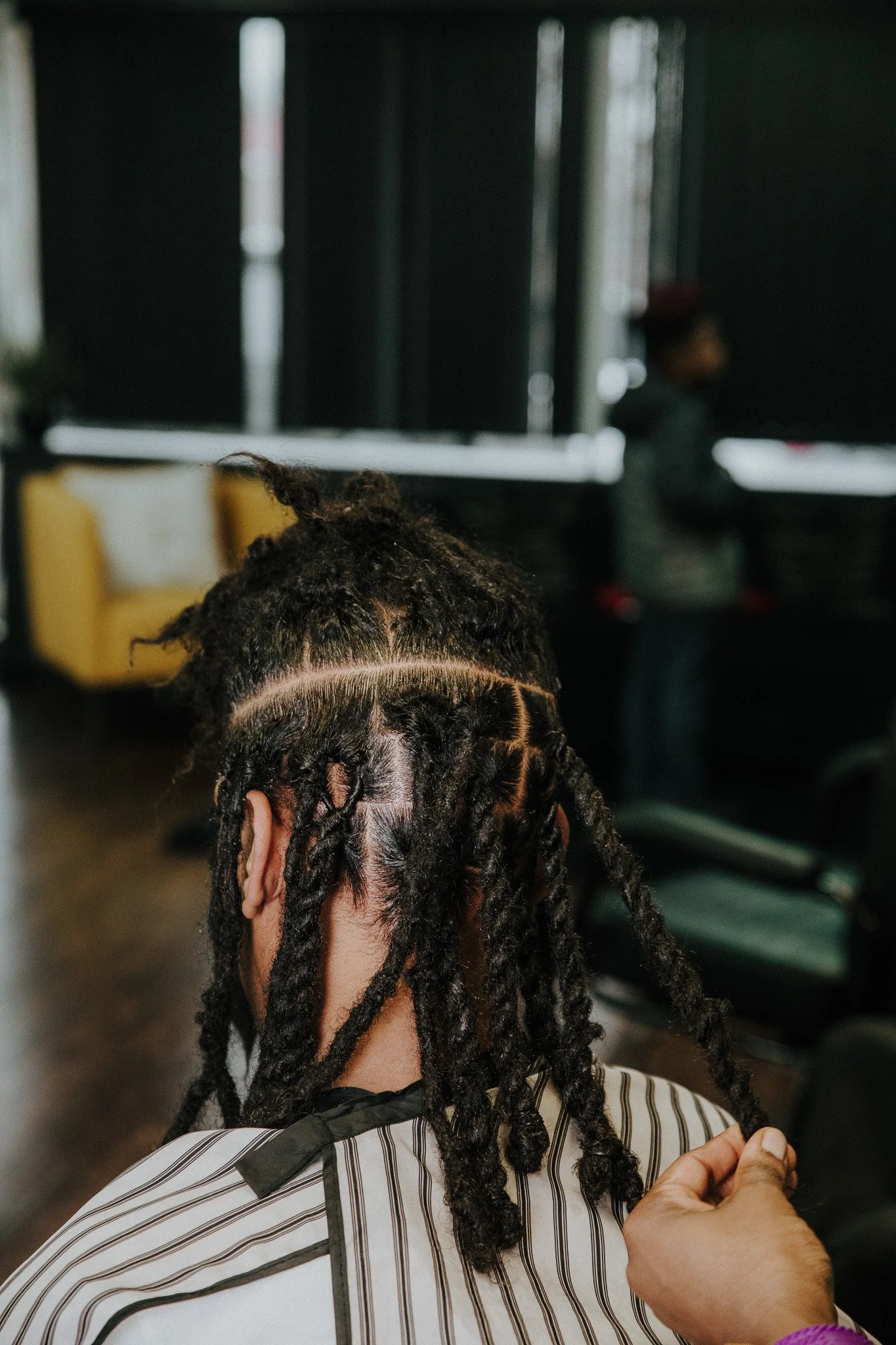 Person with freshly styled dreadlocks sitting in a salon with salon cape, and a person in the background. The dreadlocks are thick and neatly sectioned.