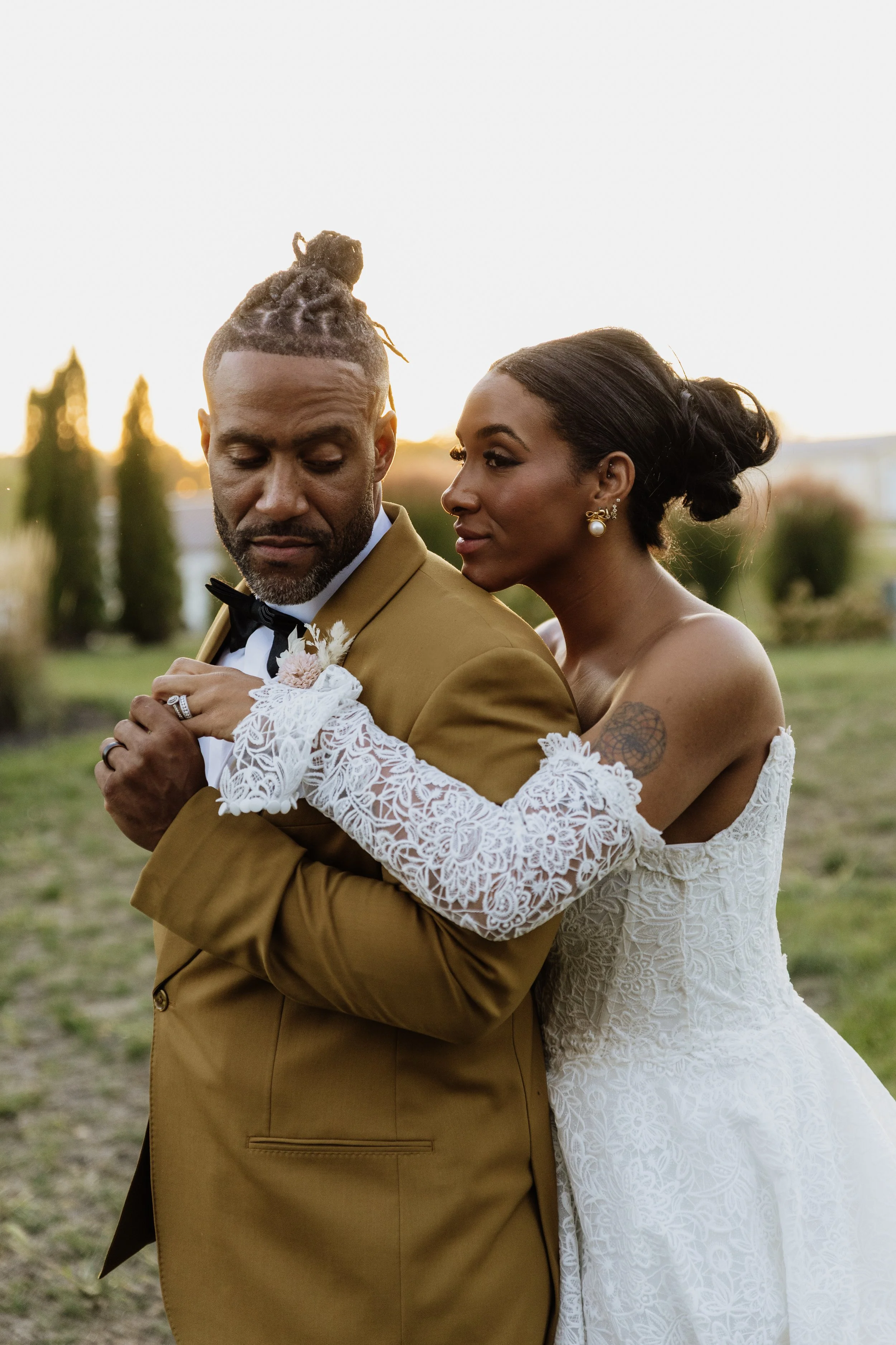 A bride and groom embrace outdoors during sunset. The groom wears a brown suit with a bow tie, while the bride wears a white lace wedding dress with lace sleeves. The bride's hair is styled in an elegant updo, and she wears pearl earrings. The groom has styled dreadlocks.