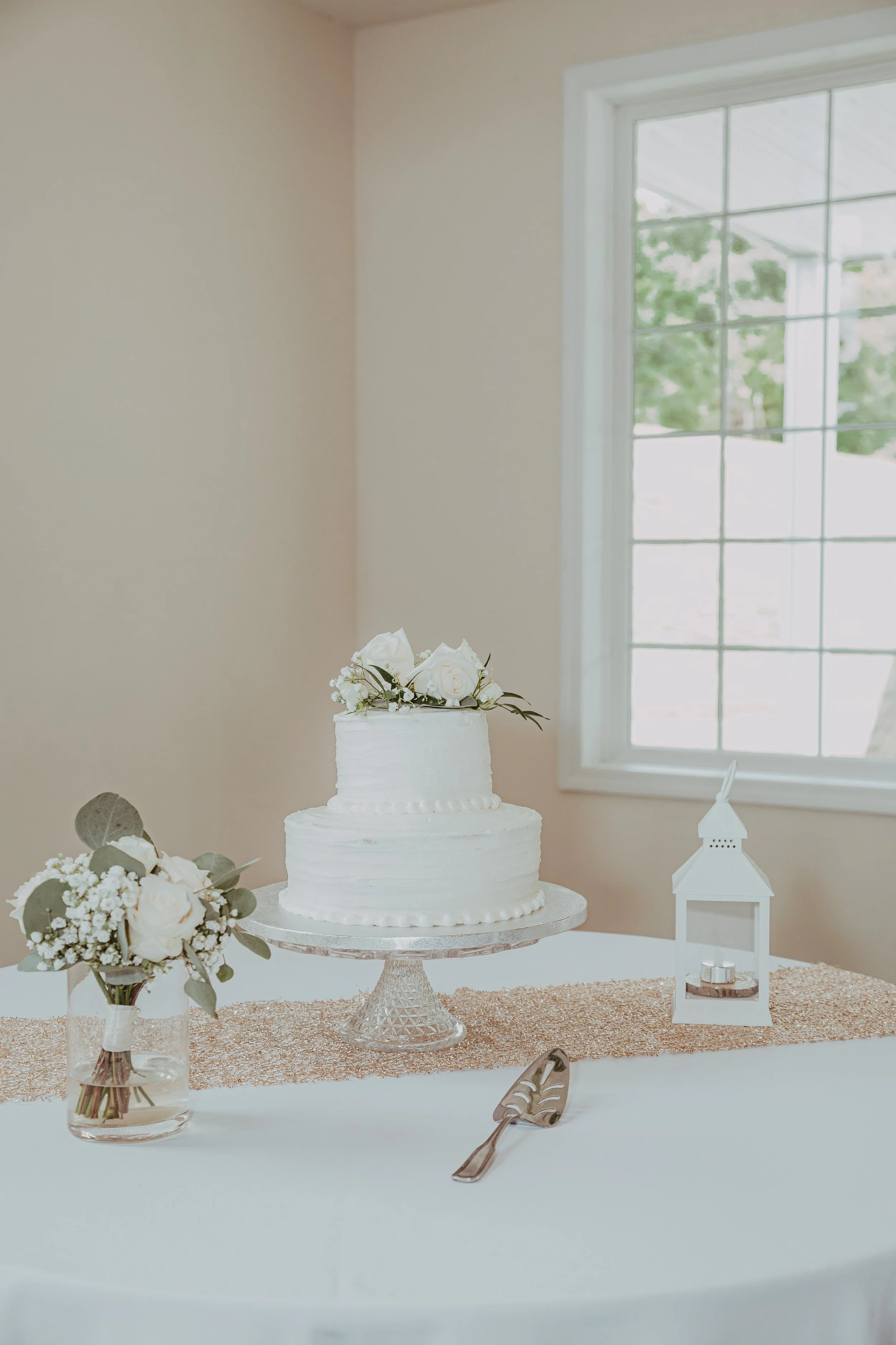 White wedding cake decorated with flowers on a table with a vase of flowers, a lantern, and a cake server, near a window.