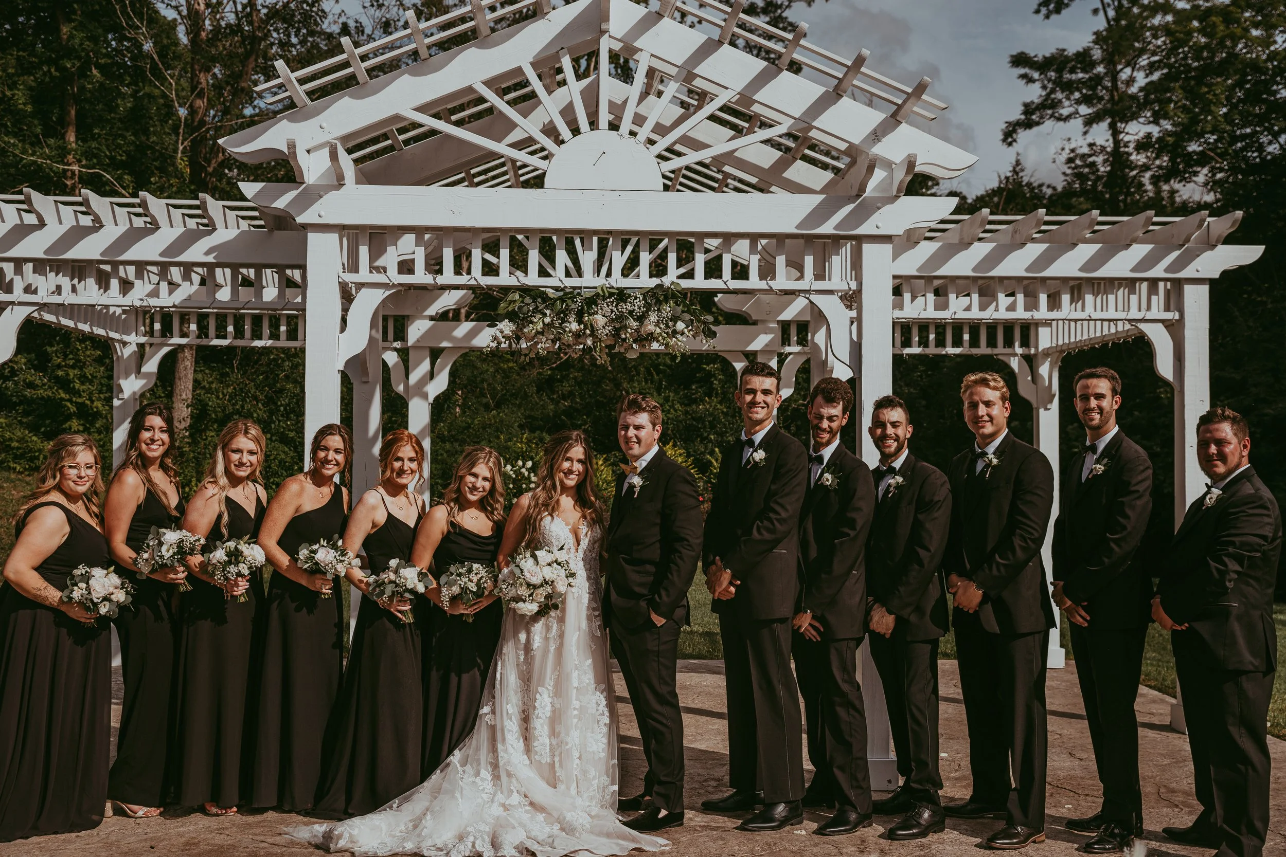 A wedding party standing outdoors in front of a white wooden arbor on a sunny day, with the bride and groom in the center, surrounded by bridesmaids and groomsmen, all smiling.
