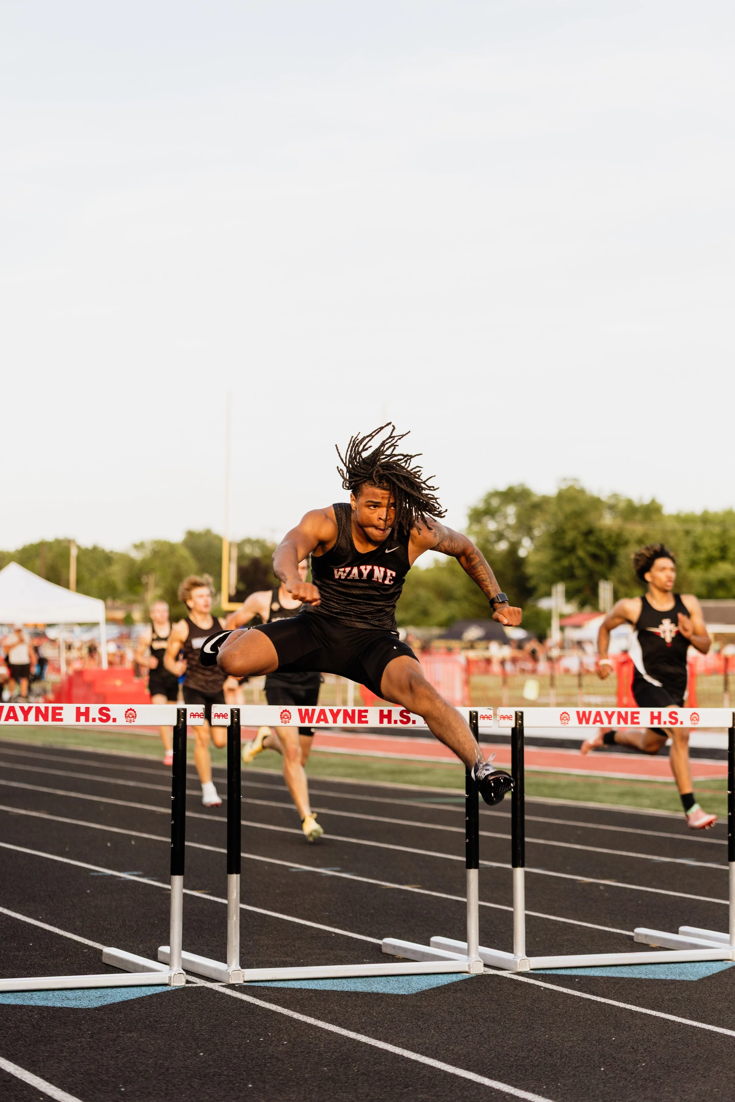 A male athlete with long hair in a black sports uniform is mid-air jumping over a hurdle during a race at Wayne High School track. Other racers and trees are visible in the background on the track field.