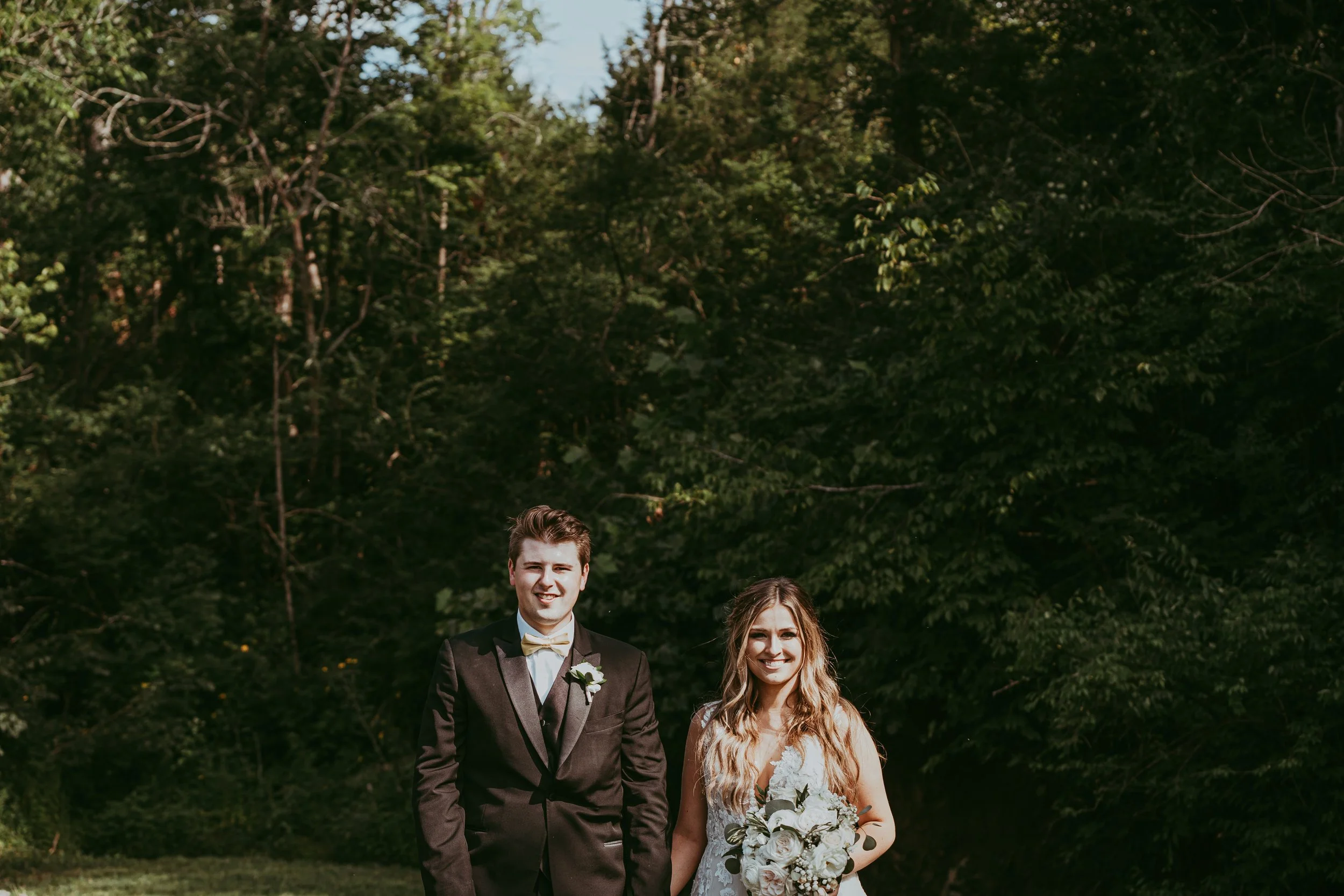A bride and groom standing outdoors in front of green trees, with the groom in a black tuxedo and the bride holding a bouquet of white flowers, both smiling.