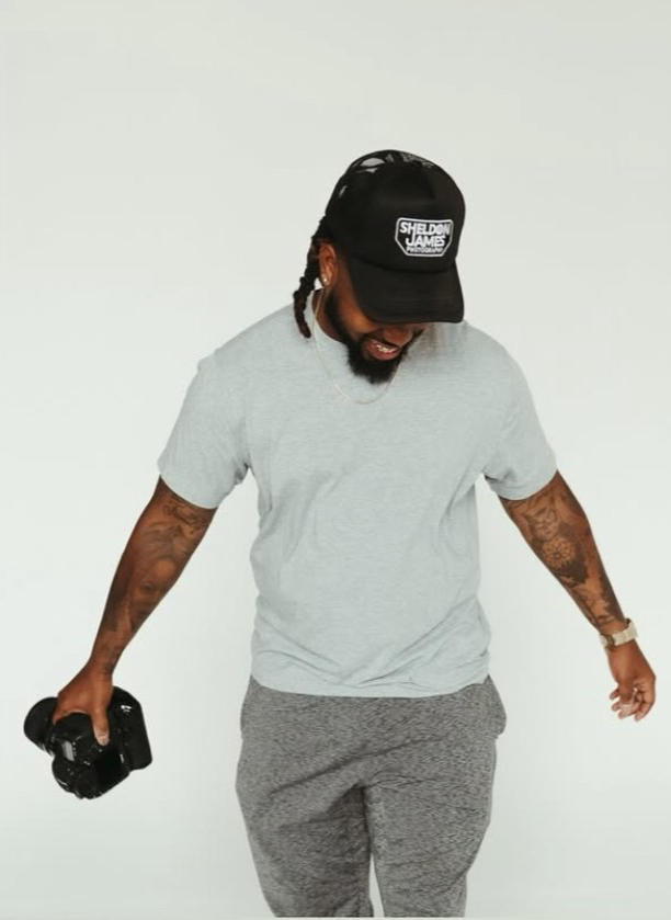 Man with tattoos, wearing a black Sheldon  James Photography cap, gray t-shirt, gray sweatpants, holding a camera, smiling, in a plain white background.