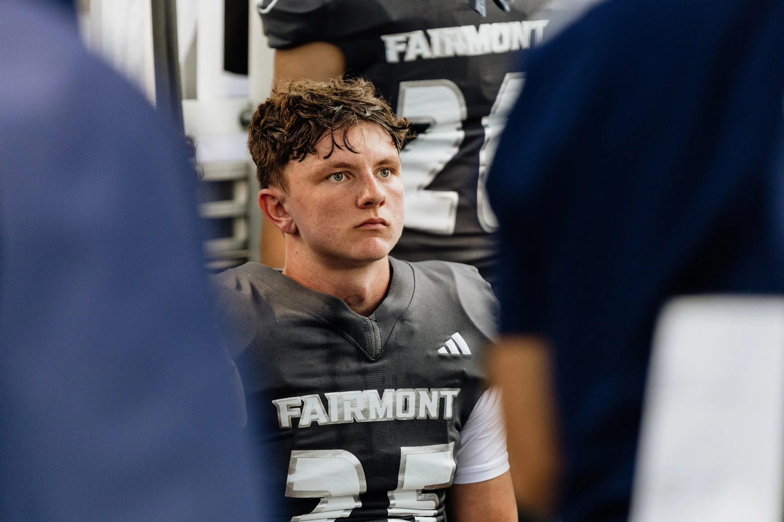 A young football player with curly brown hair and light skin, wearing a dark gray and white football uniform with 'FAIRMONT' written on the chest, sits on the sideline with a serious expression, surrounded by teammates and coaches.