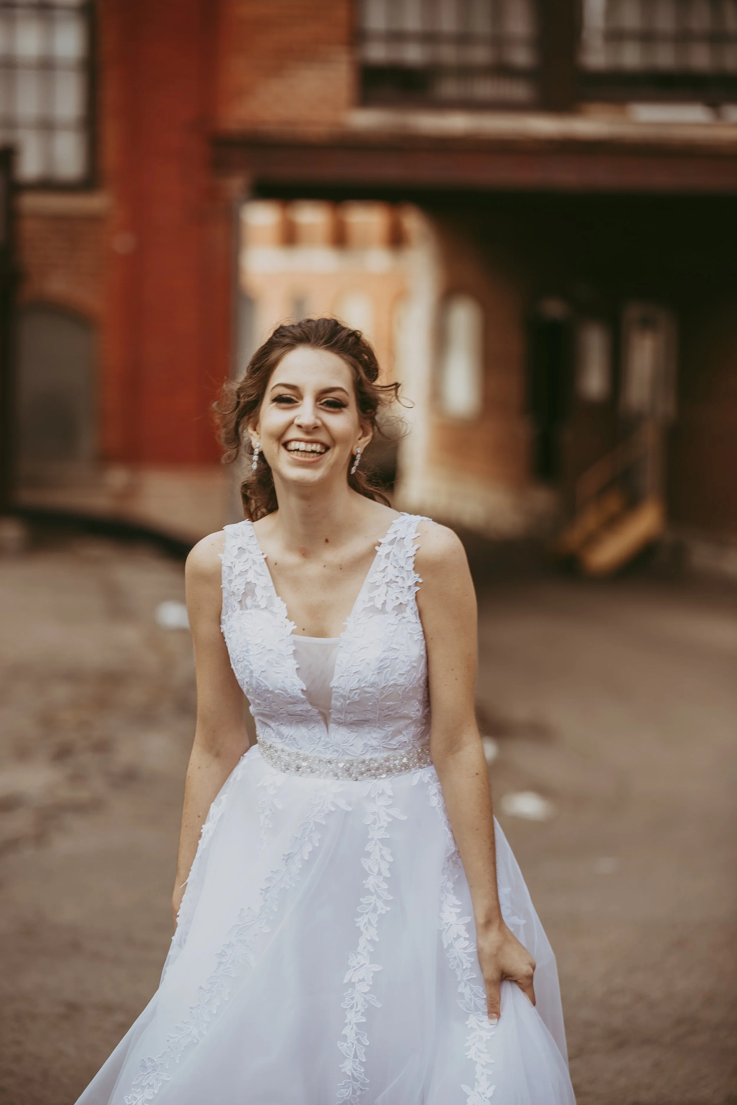 A woman in a white wedding dress smiling outdoors with a blurred brick building in the background.