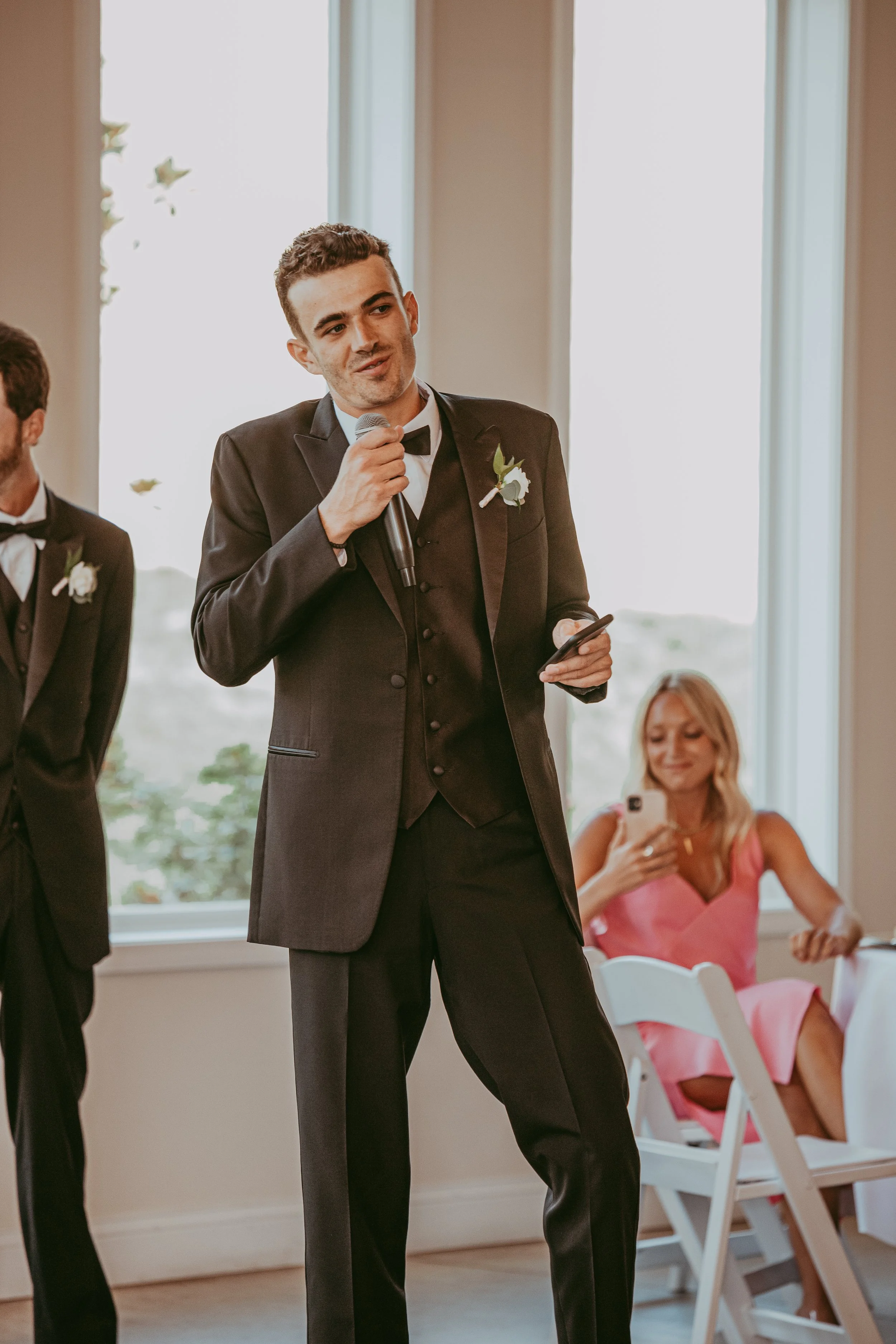 A groom in a black tuxedo with a bow tie, holding a microphone and a phone, giving a speech at a wedding reception. A woman in a pink dress is sitting behind him, smiling and looking at her phone.