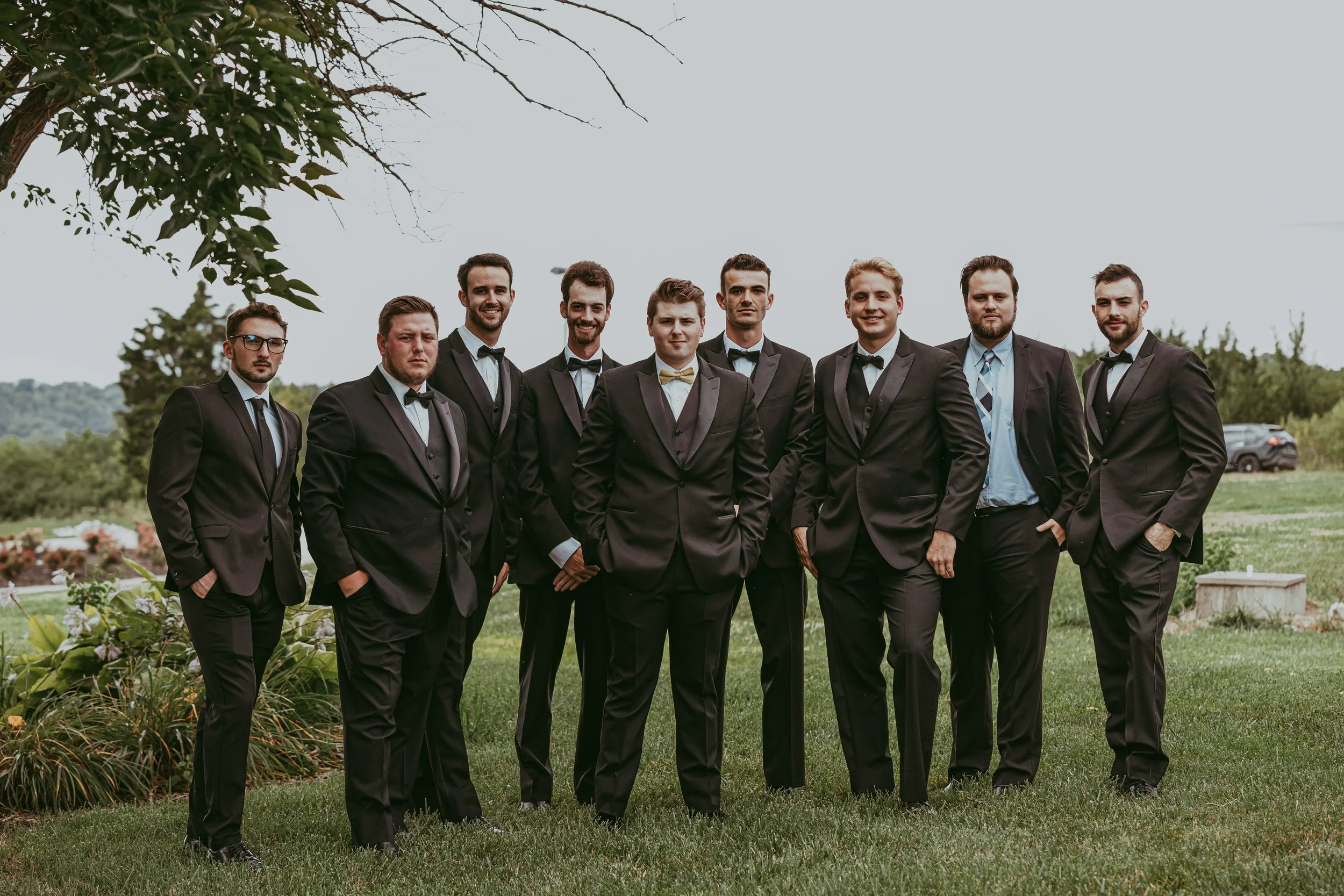 A group of ten men in black tuxedos with ties and bow ties standing outdoors on green grass with trees and hills in the background.