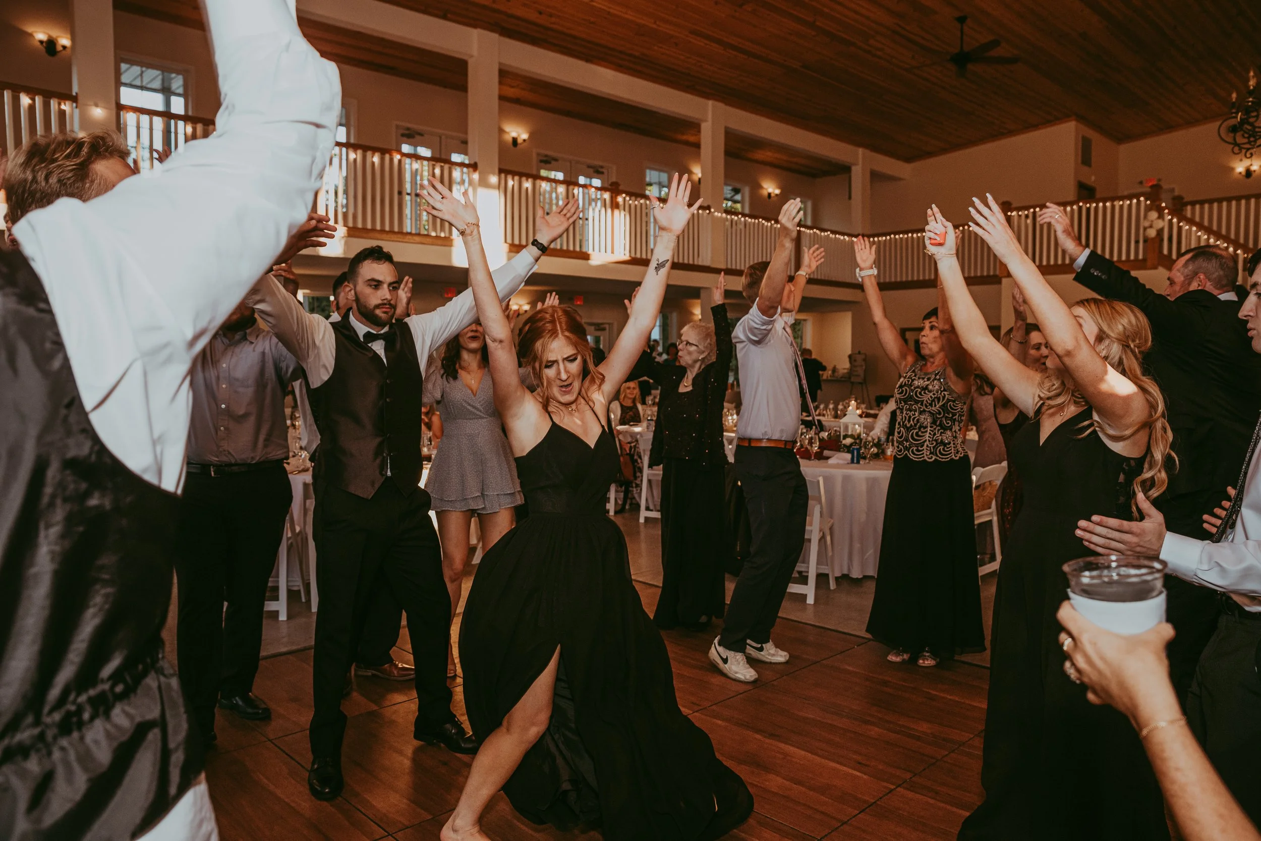 People dancing and celebrating at a wedding reception with their hands raised in the air, inside a decorated hall with wooden ceiling and string lights.