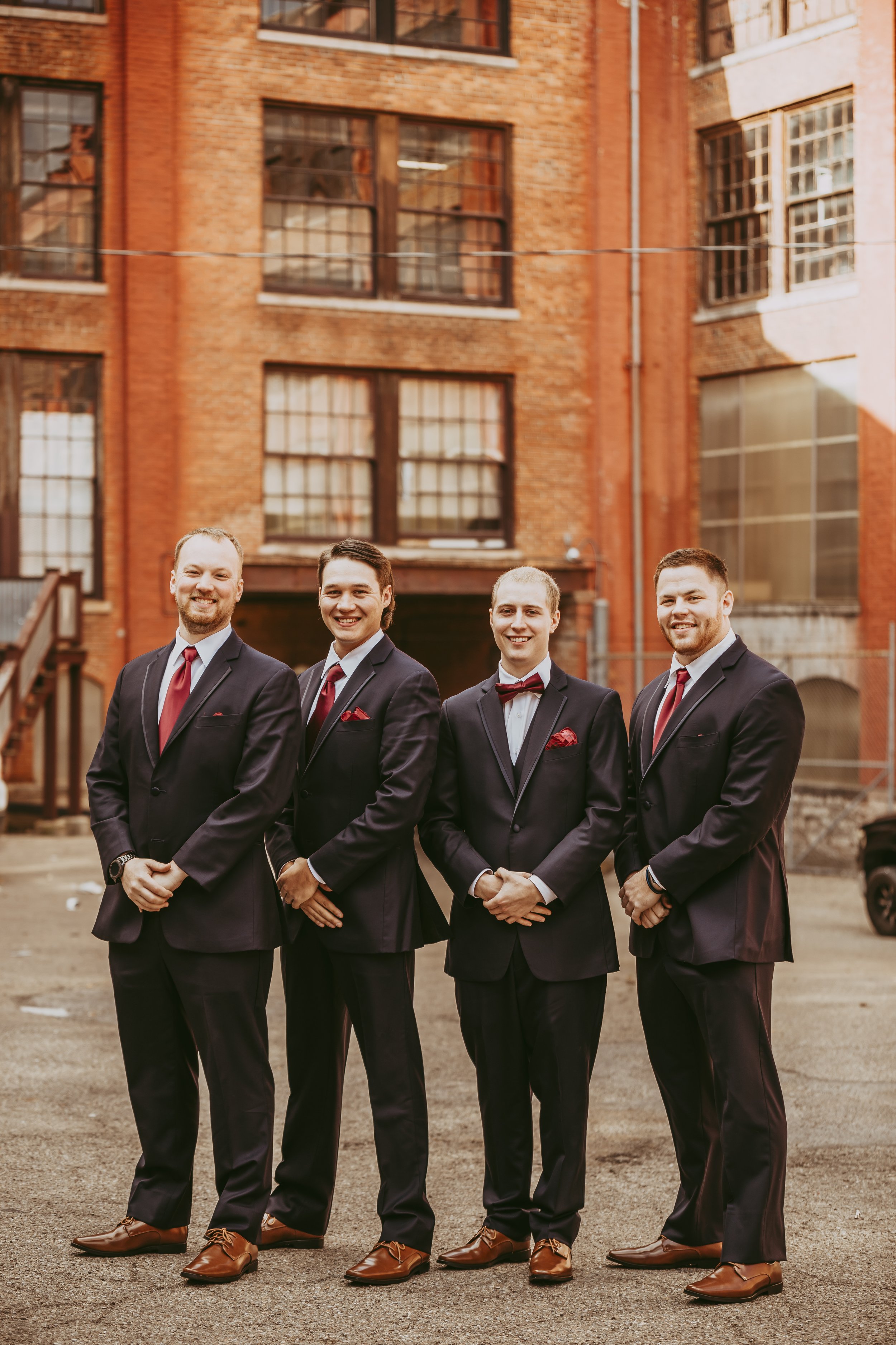 Four men in black suits with red ties and pocket squares standing outside in a parking lot, in front of a brick building with large windows.