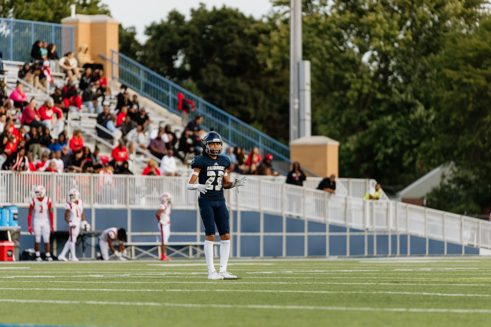 Football player in uniform standing on the field during a game, with spectators seated in the stands behind.