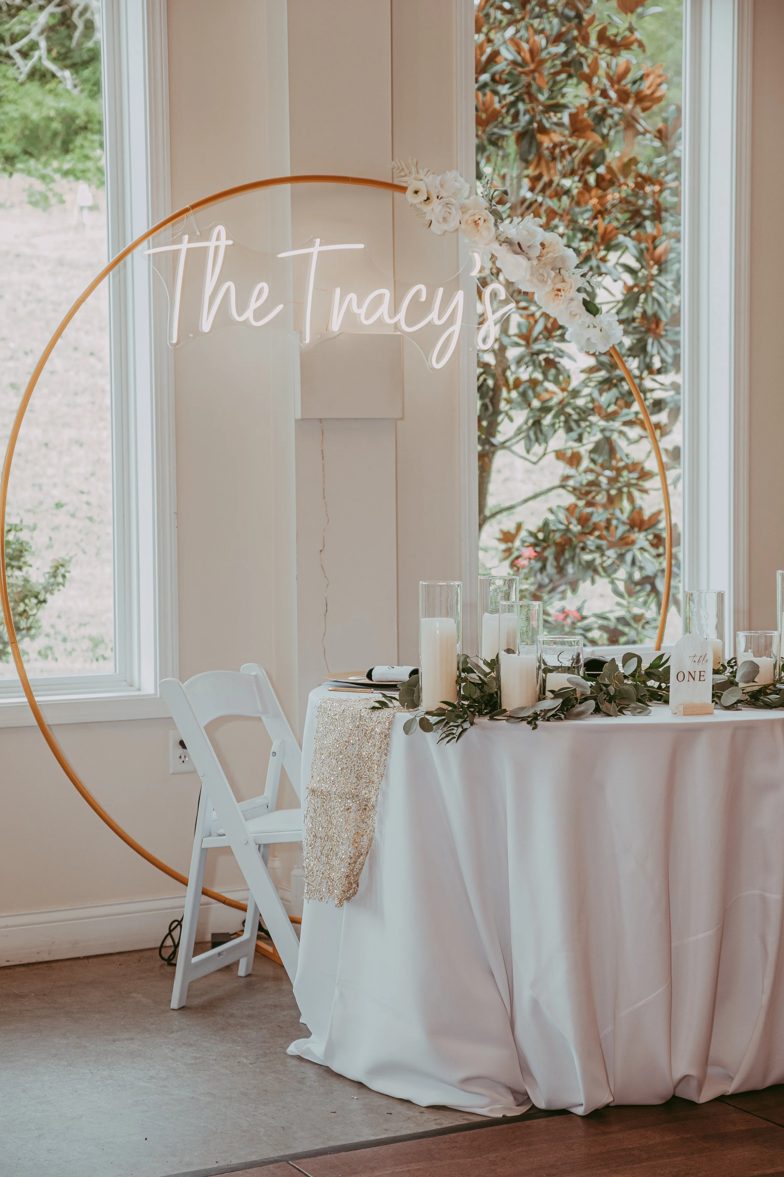 Decorated sweetheart table with white tablecloth, candles, greenery, and an illuminated neon sign saying 'The Tracys' behind the table at a wedding reception.