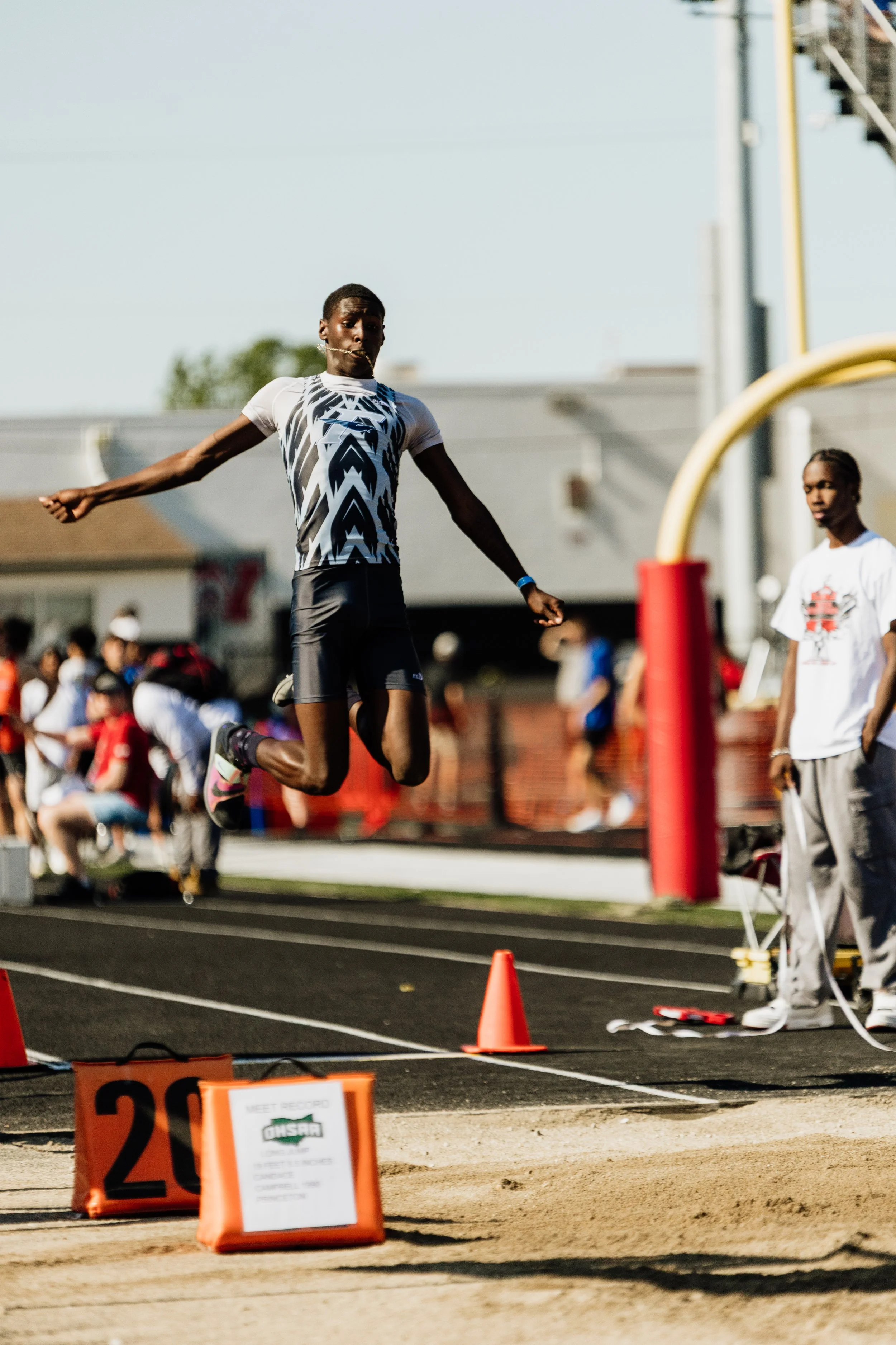 A male athlete in mid-air jumping over a hurdle during a track and field event at an outdoor stadium with other athletes and spectators in the background.