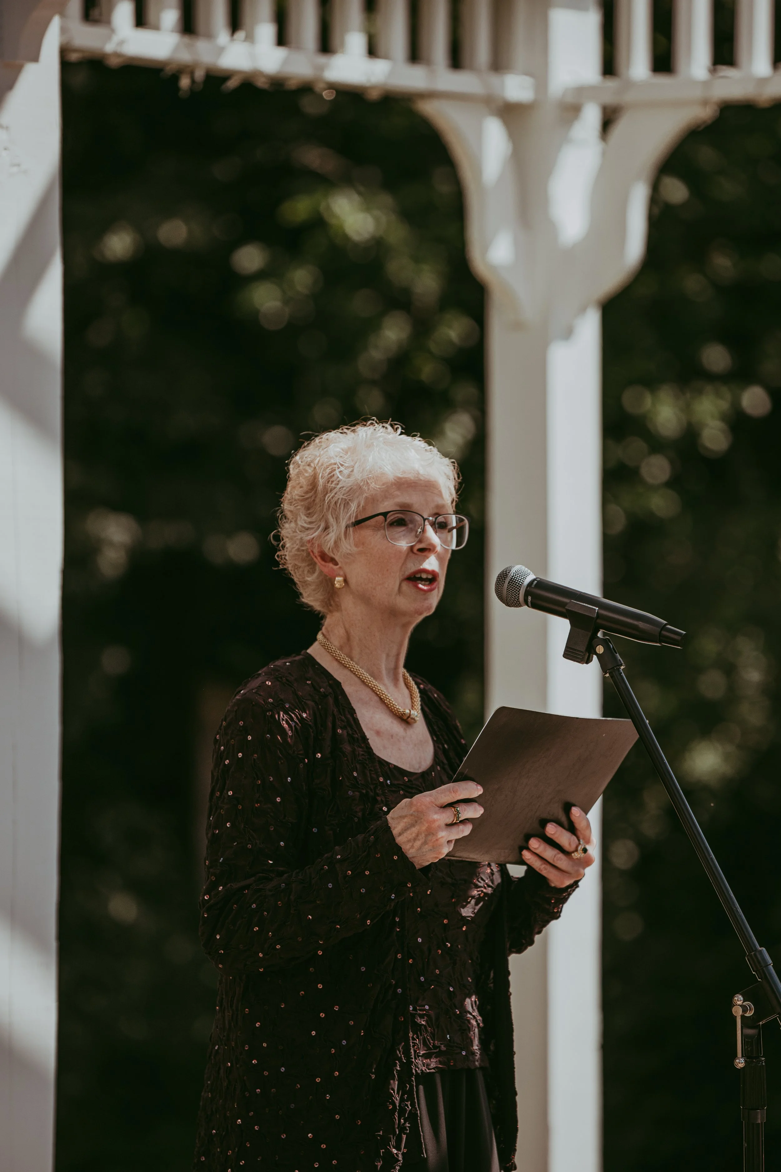 An elderly woman with white hair, glasses, and a pearl necklace speaking into a microphone outdoors, holding a piece of paper.