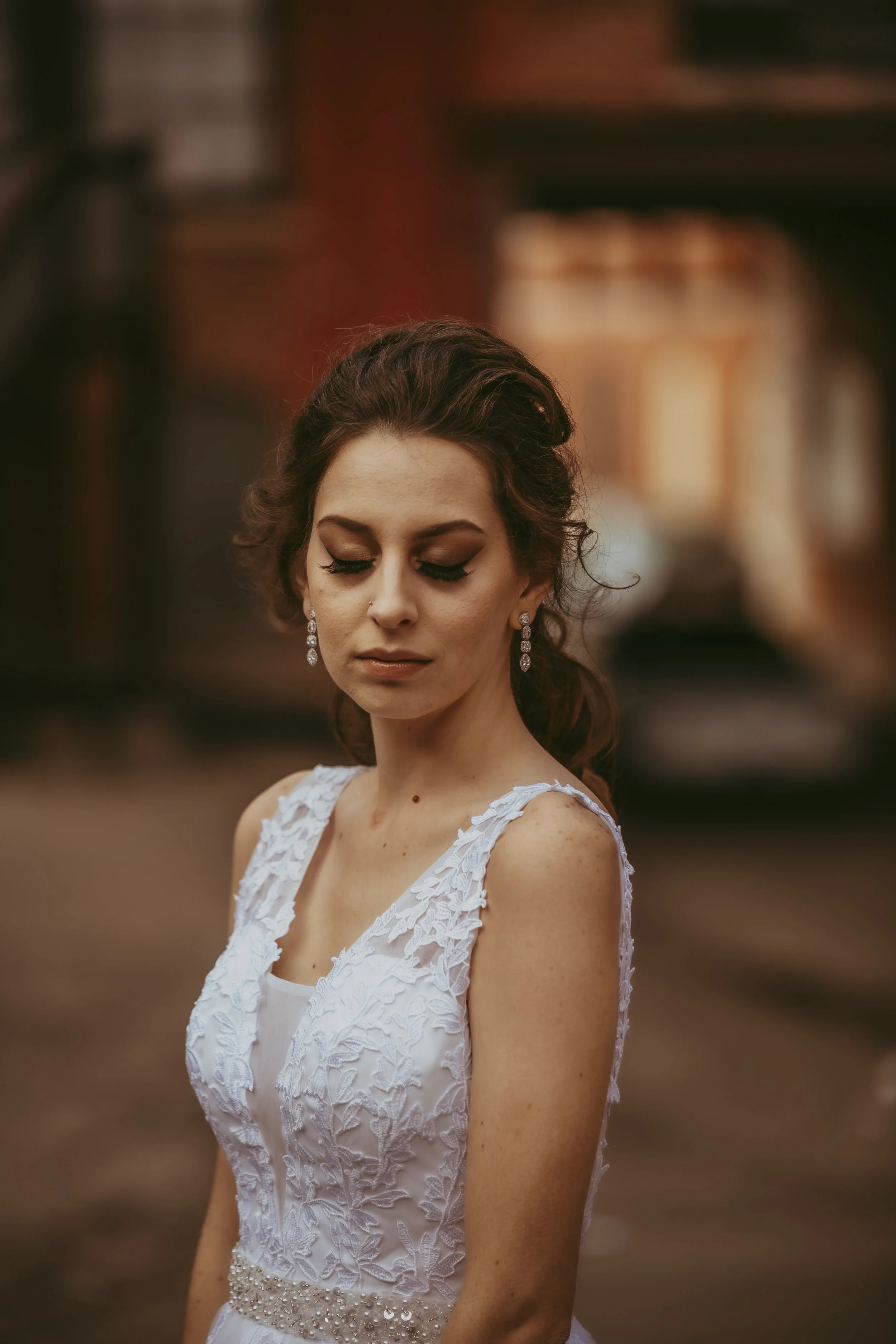 A bride with dark, wavy hair styled back, wearing a sleeveless white lace wedding dress with floral details and a beaded waistband, along with dangling earrings. She has a serene expression with closed eyes, standing outdoors with a blurred backgroun