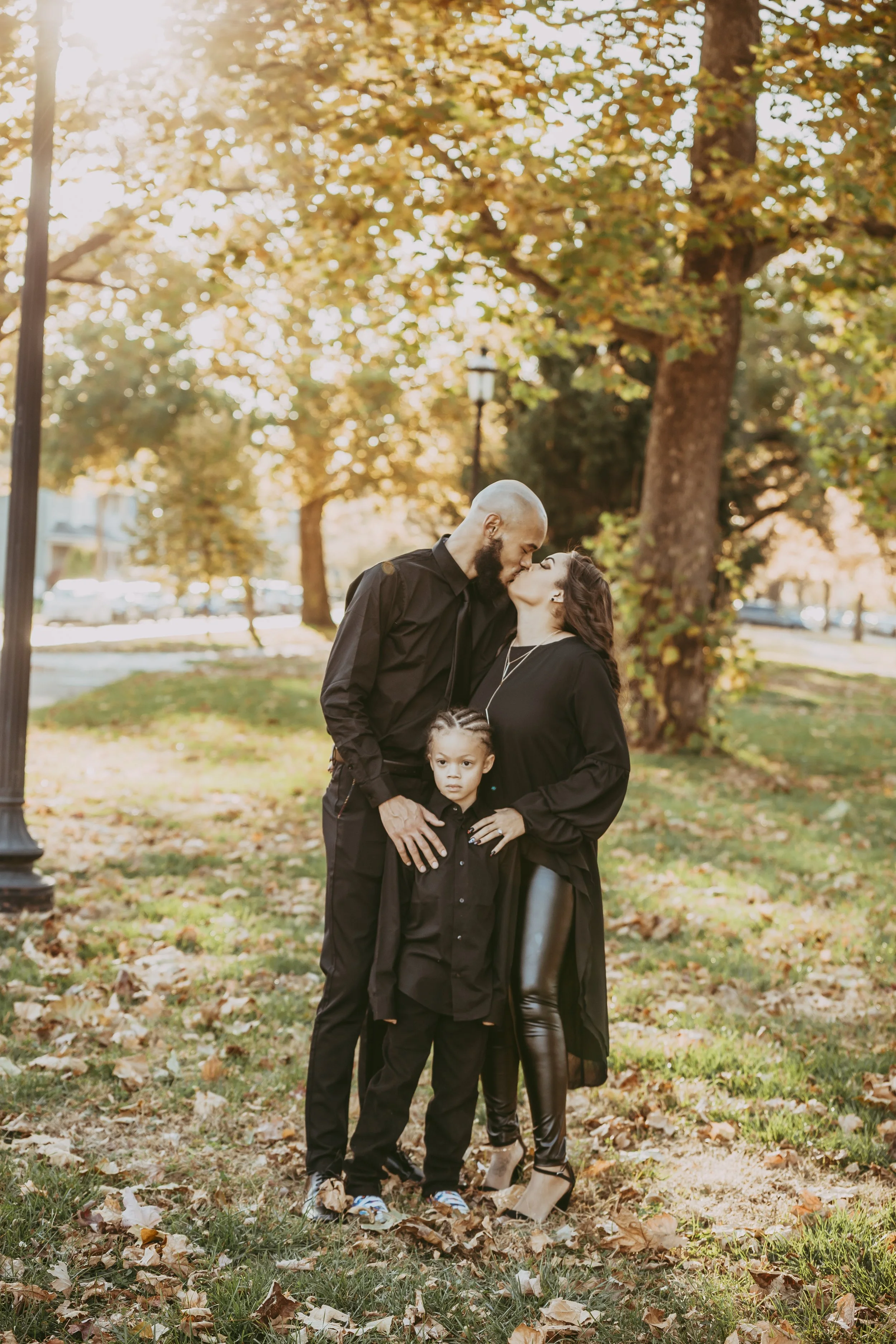 A family of three, a man, a woman, and a young girl, standing outdoors on a fall day, with sunlit trees and fallen leaves in the background. The man and woman are sharing a kiss, while the girl looks at the camera.