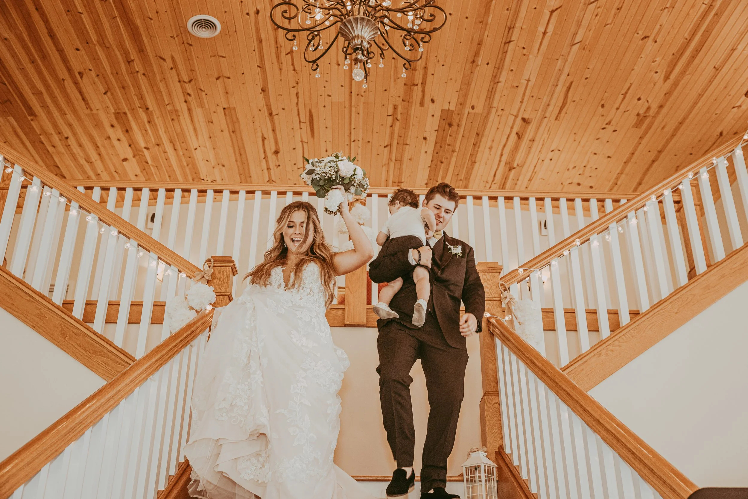 A bride and groom smiling, celebrating their wedding on a staircase, holding a young child. The bride is in a white lace wedding dress, and the groom is in a dark suit. The bride holds a bouquet of flowers.
